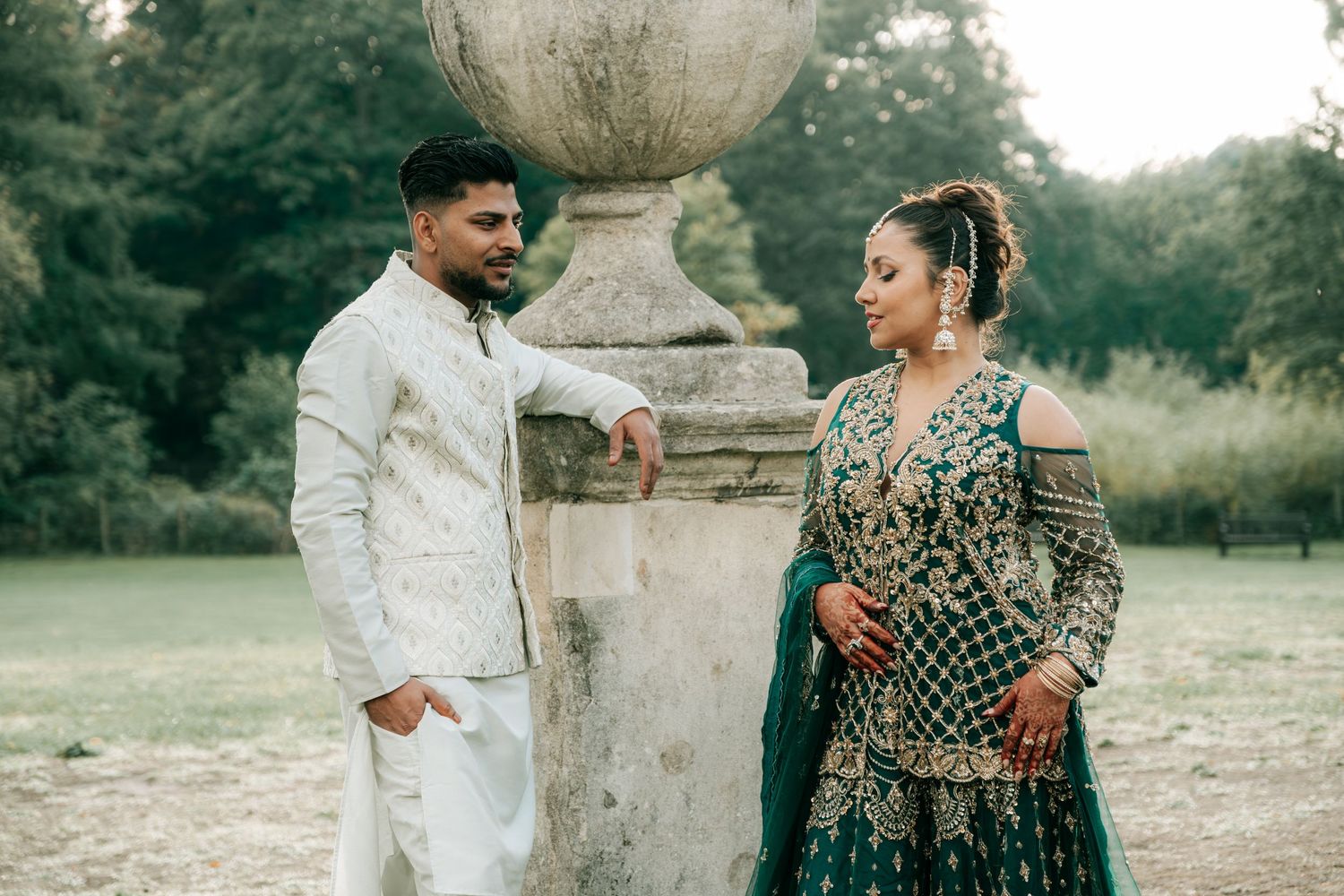 A couple in traditional wedding attire shares a moment near a decorative stone pillar in a garden setting.