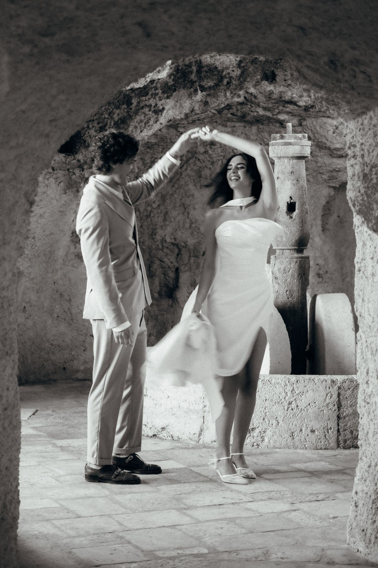 Black and white photo of a couple dancing in a rustic cave setting, groom spinning bride in white dress.
