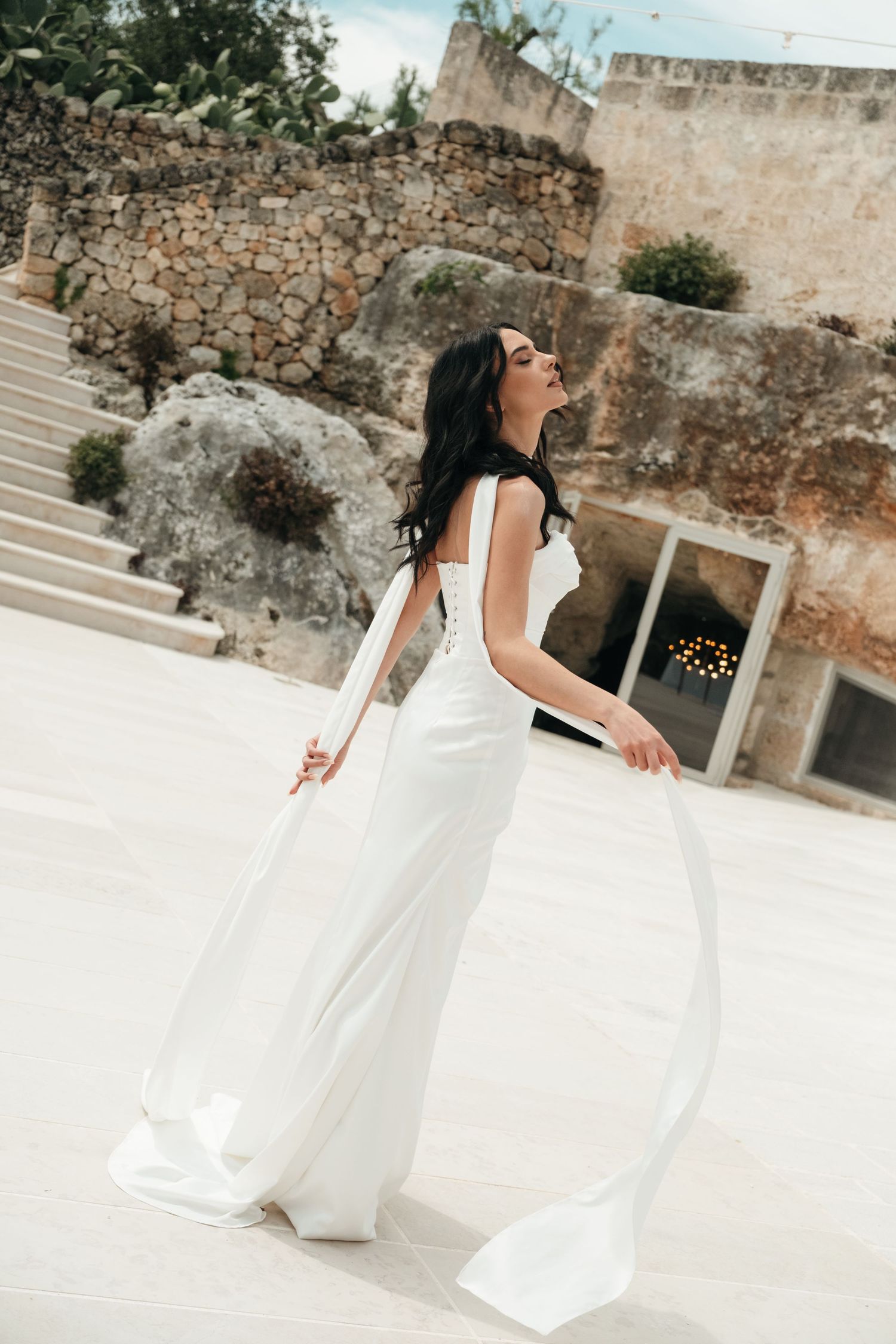 Elegant bride in white gown with flowing cape twirling outdoors at a stone venue.