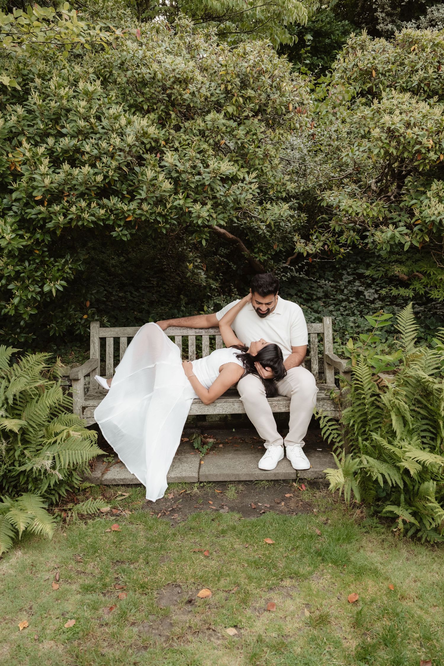 A couple in coordinated white outfits sits together on a garden bench surrounded by greenery.
