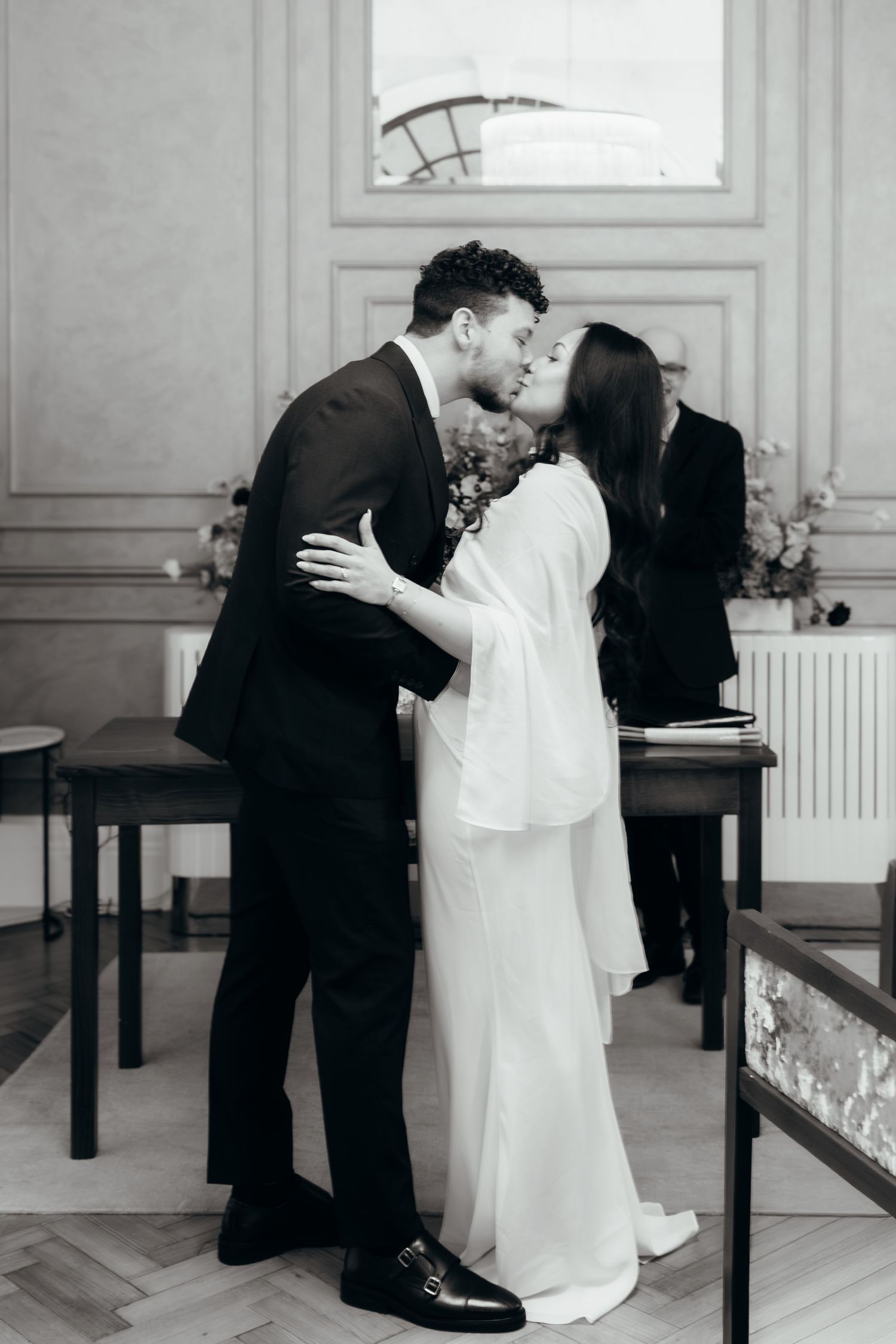 Black and white photo of a bride and groom sharing their first kiss at a civil wedding ceremony indoors.
