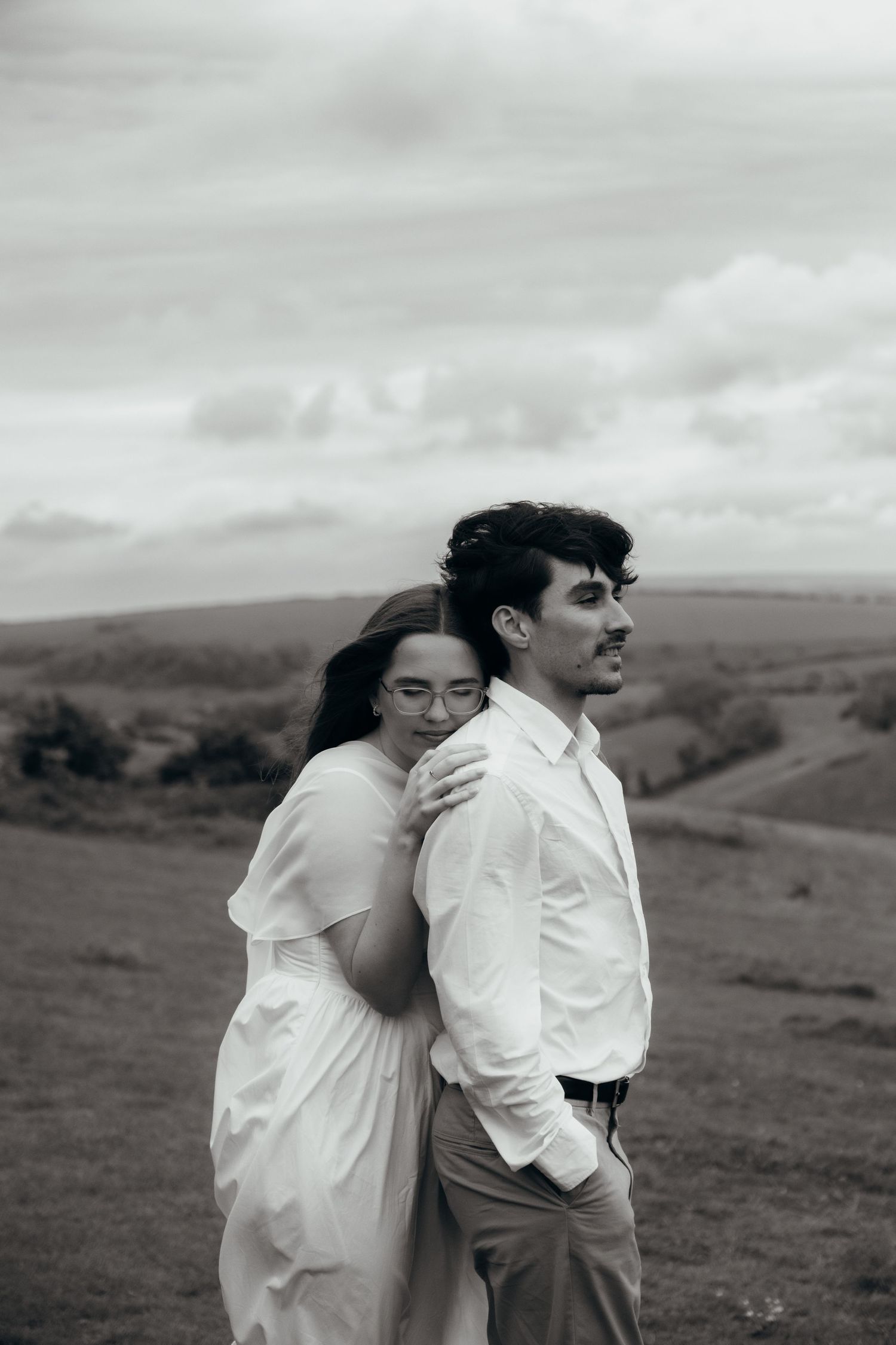 Black and white photo of a couple in a field, woman in white dress embracing man in white shirt under cloudy sky.