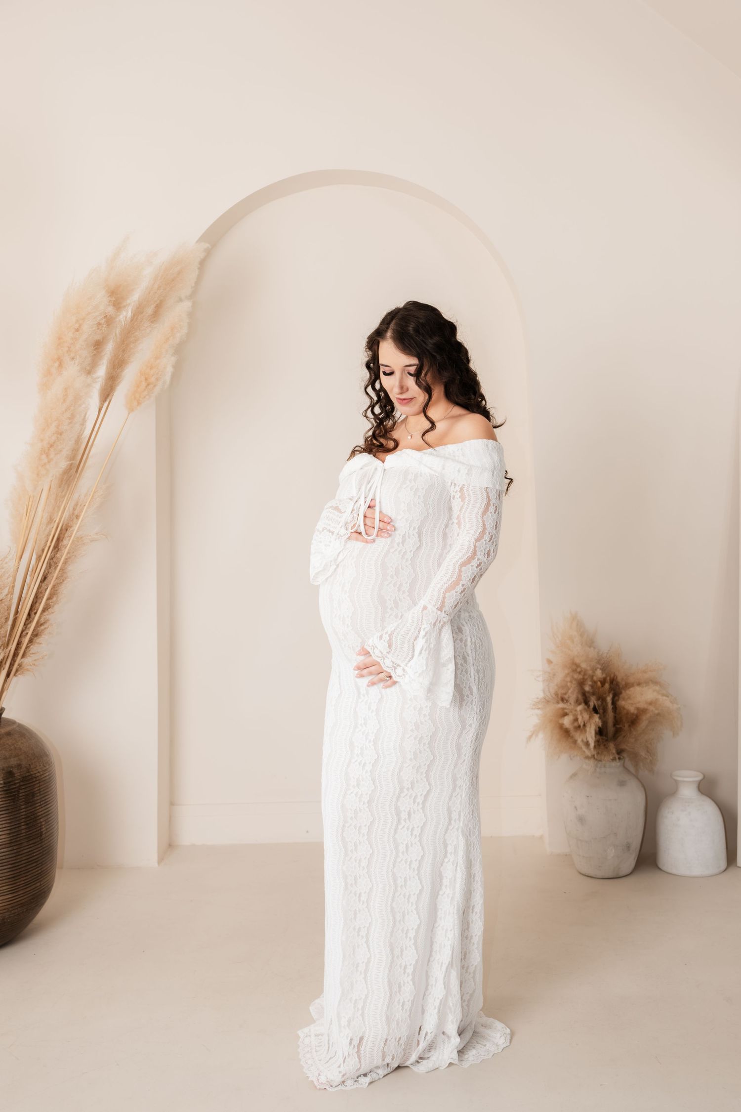 A dreamy maternity photo in a white lace dress against a neutral backdrop with pampas grass decor.