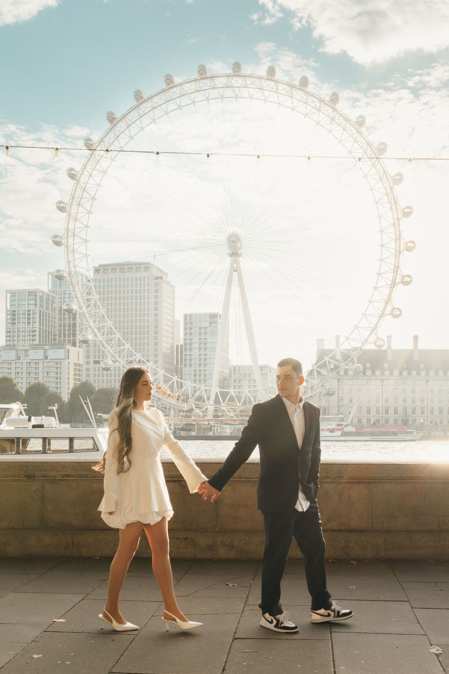 A couple takes romantic photos with the London Eye ferris wheel visible in the background at sunset.
