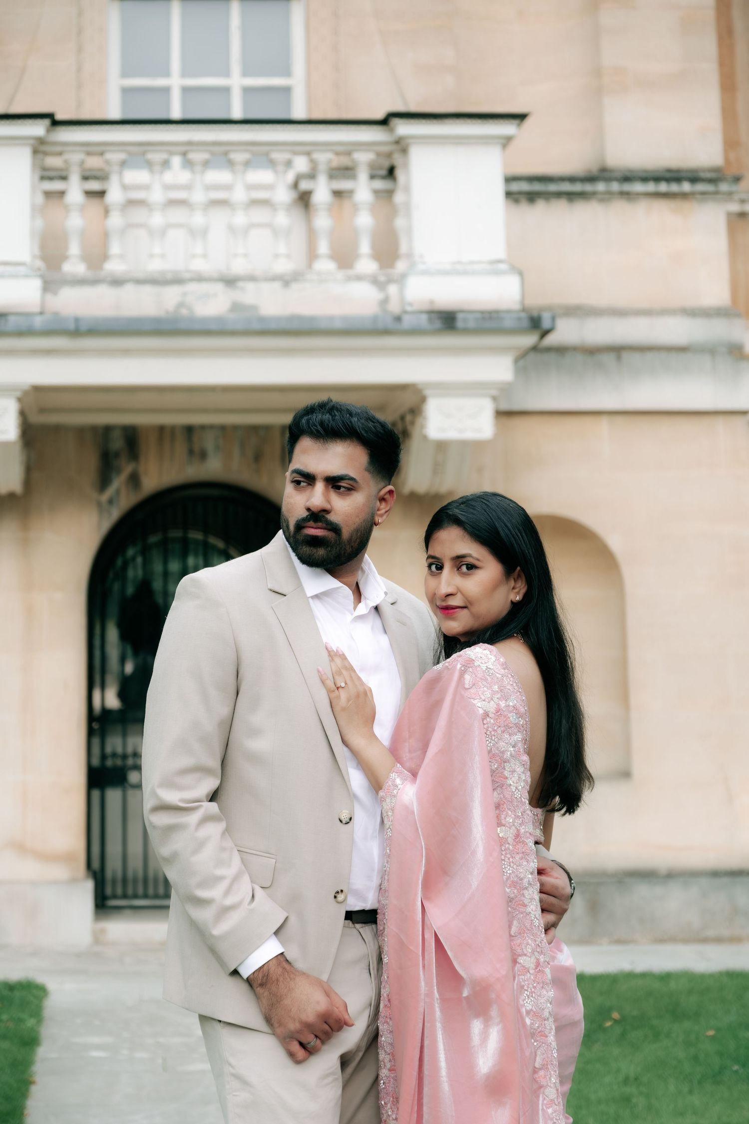 A couple in traditional South Asian attire poses elegantly in front of a classical building.