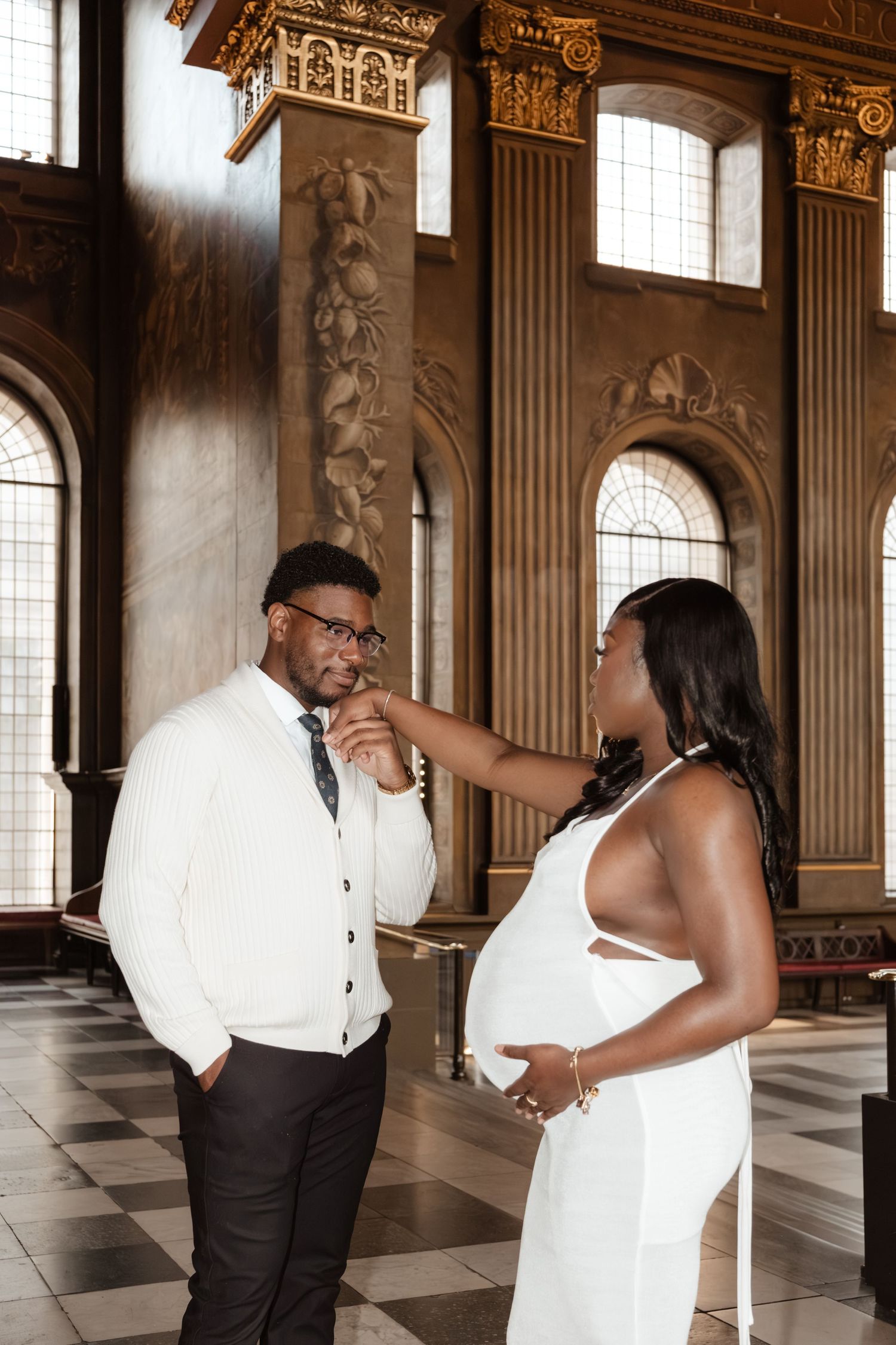 A couple in formal attire shares playful moments in an elegant hall with ornate architecture and arched windows.