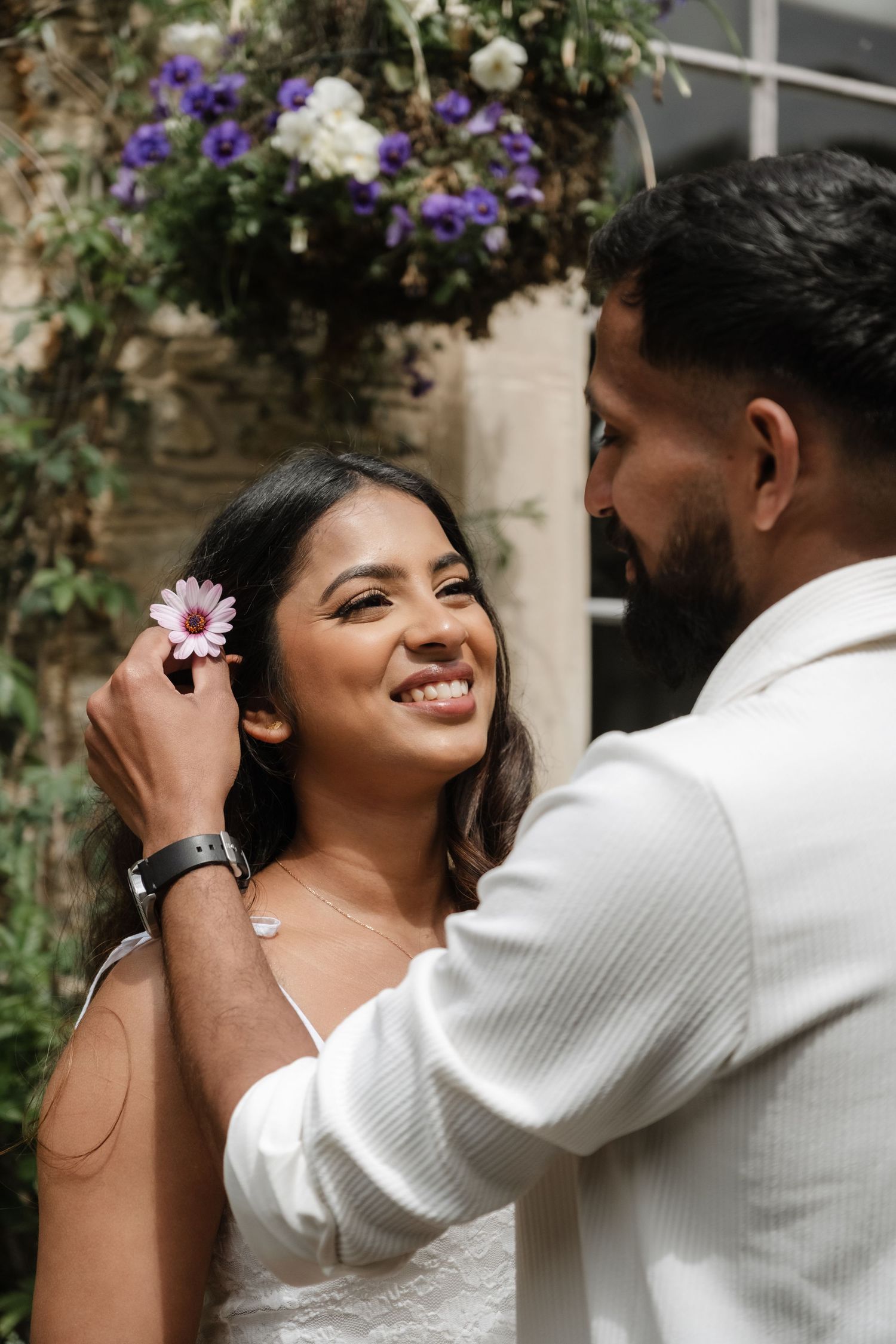 Groom placing a pink flower in bride's hair, both smiling, with purple and white floral arch backdrop.