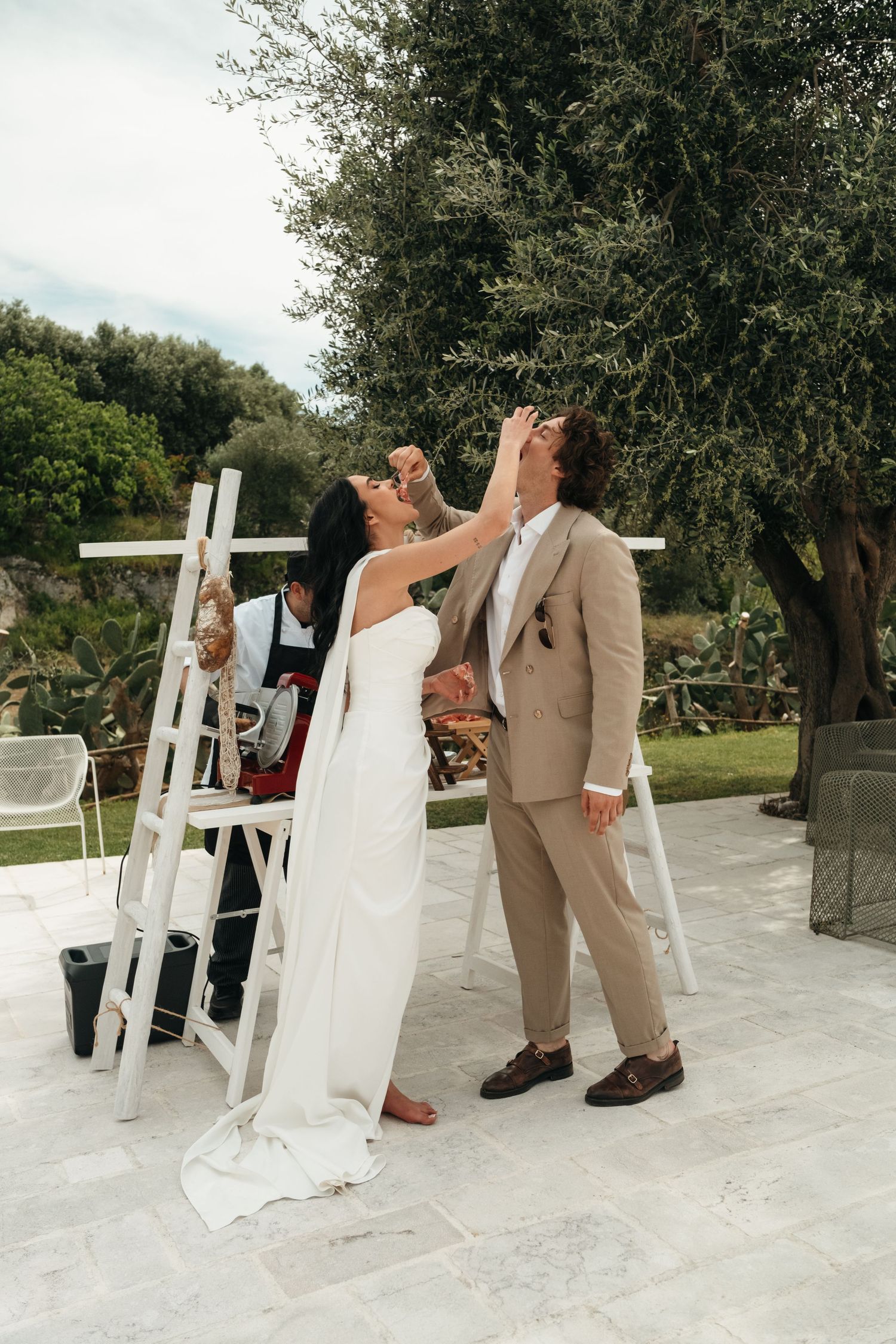 Bride and groom sharing a joyful moment outdoors at their wedding ceremony, with white ladders and olive trees in background.