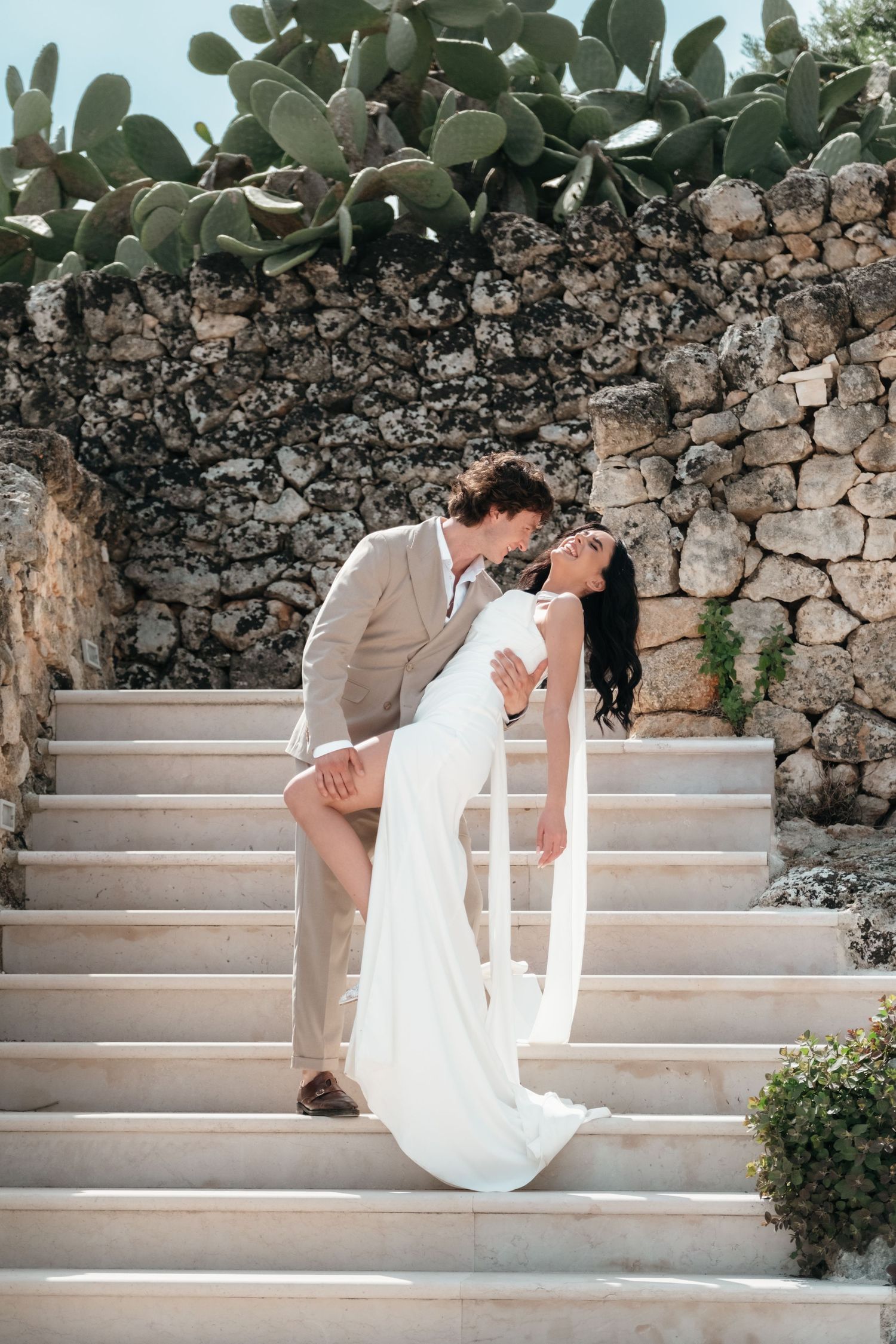A bride and groom share a romantic embrace on stone steps, with cacti and a rustic wall backdrop.