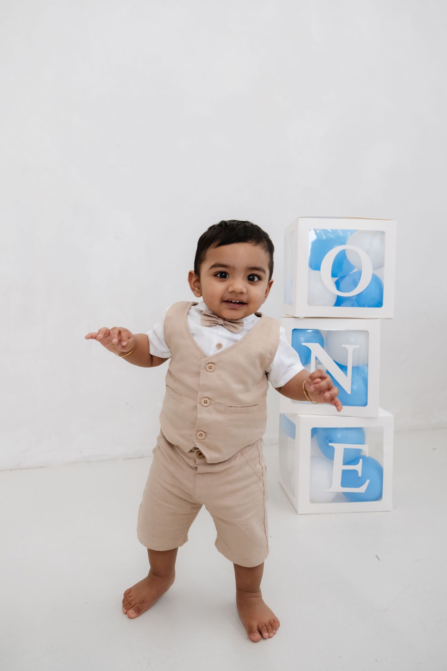 Adorable baby boy in beige vest outfit standing next to stacked ONE blocks celebrating his first birthday.
