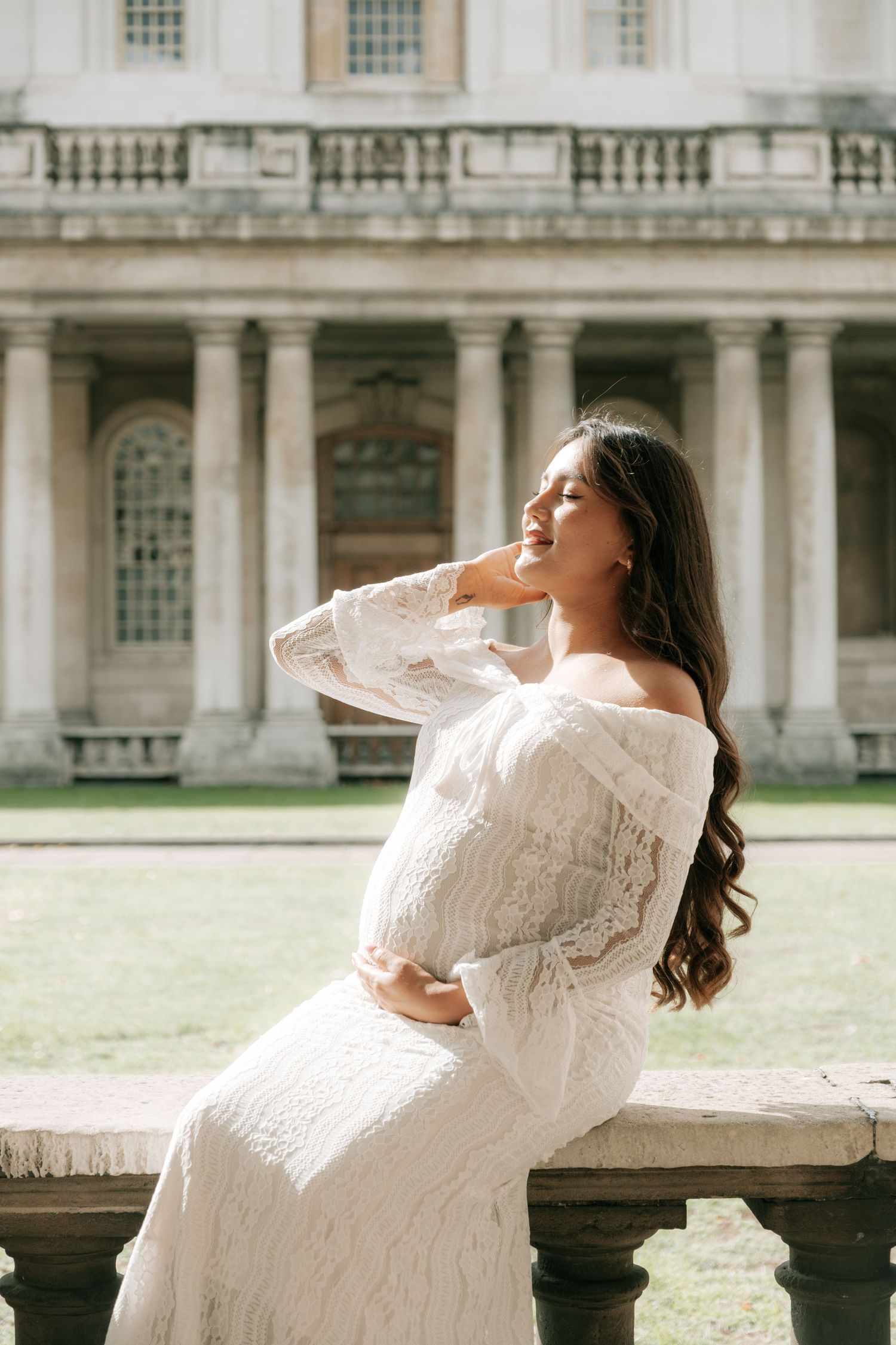 A woman in a white lace dress poses gracefully against a historic stone building backdrop in an elegant photoshoot.