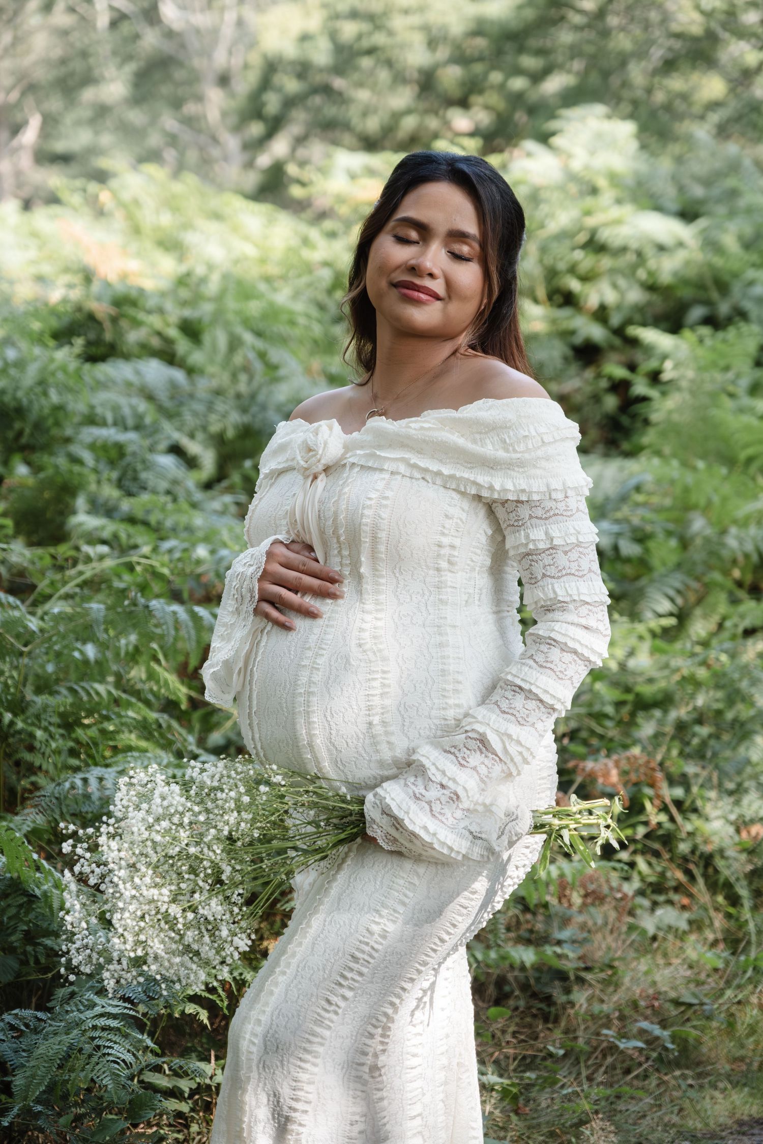 Elegant maternity portraits featuring white lace dress and baby's breath flowers in forest setting.