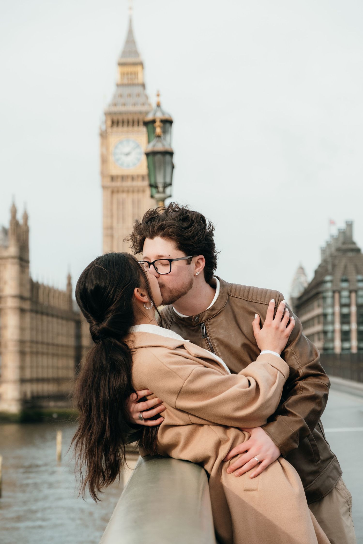 Couple kissing romantically on Westminster Bridge with Big Ben and Houses of Parliament in London background.