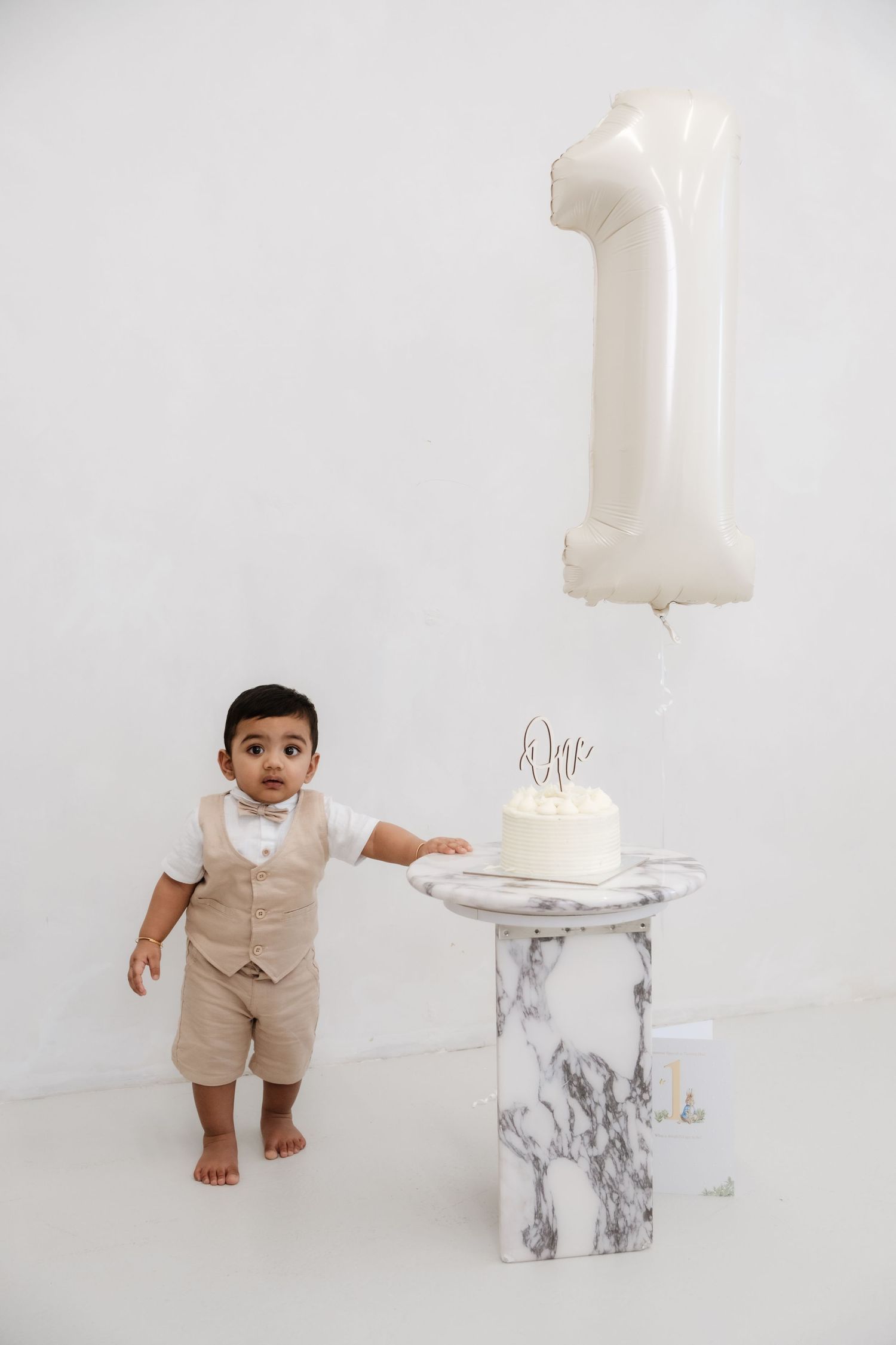 Baby boy in beige outfit touching first birthday cake beside a number 1 balloon on marble pedestal.