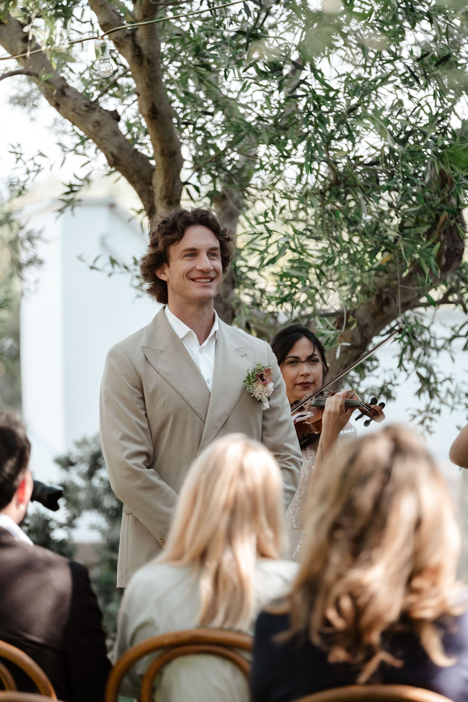 Smiling groom in beige suit with boutonniere stands at outdoor wedding ceremony while violinist performs behind him.