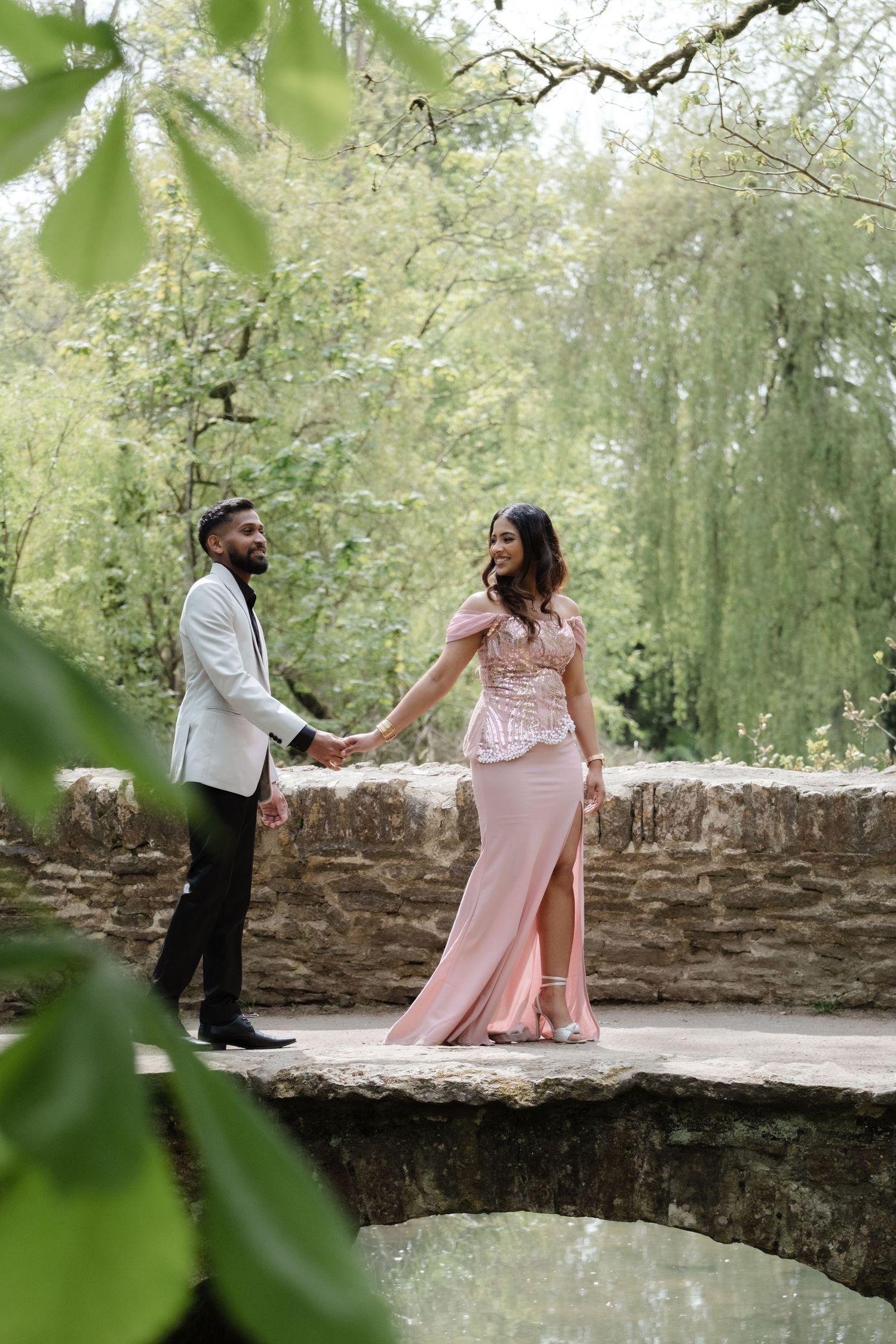 Couple in formal wear holding hands on stone bridge surrounded by lush green trees in a garden setting.