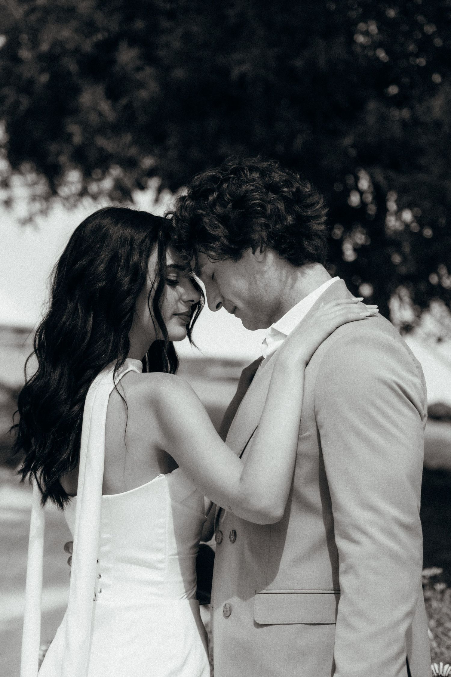 Black and white photo of a bride and groom in an intimate moment, foreheads touching, outdoors near trees.