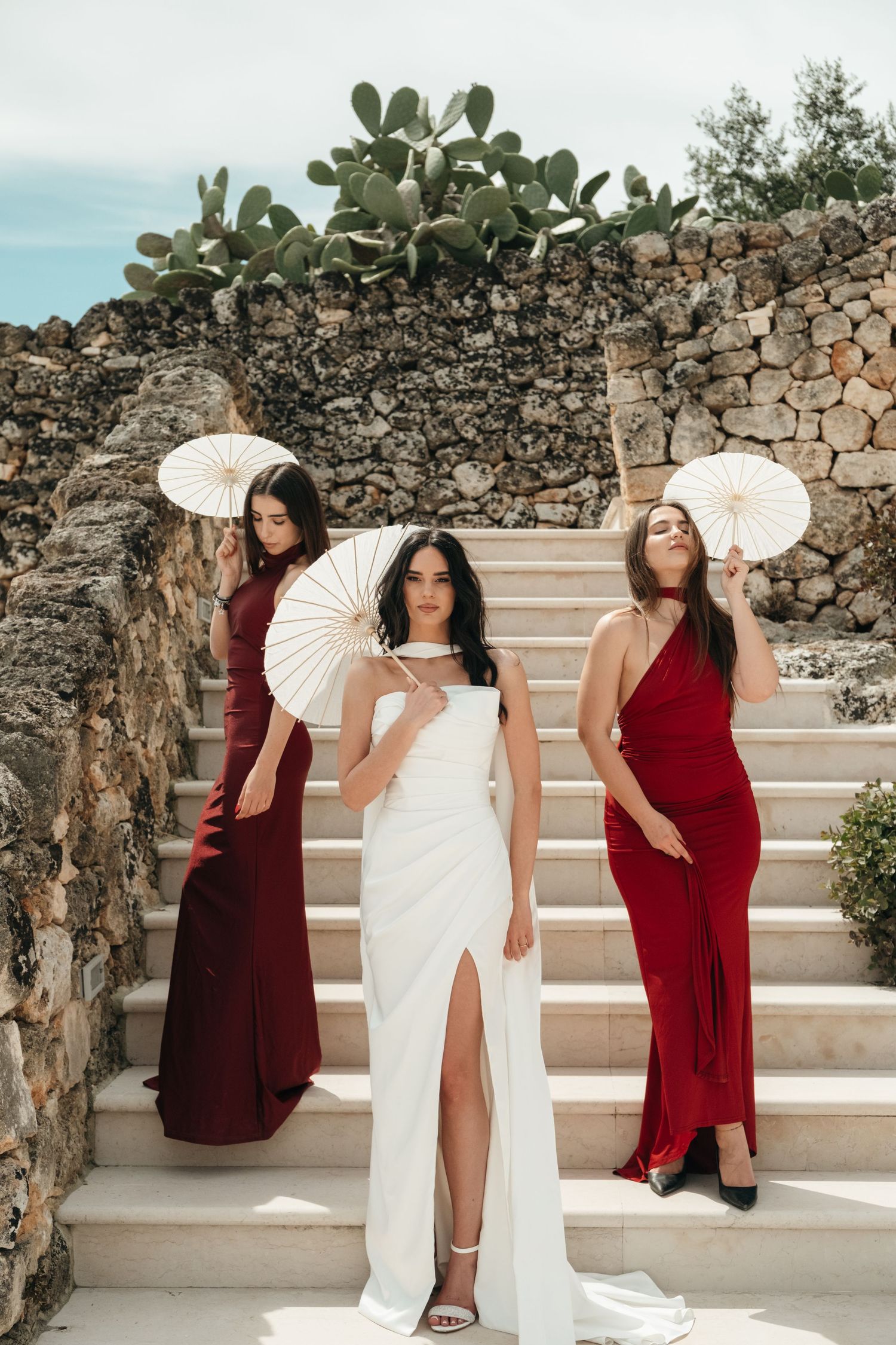 Bride in white dress poses with two bridesmaids in red gowns holding parasols on stone steps outdoors.