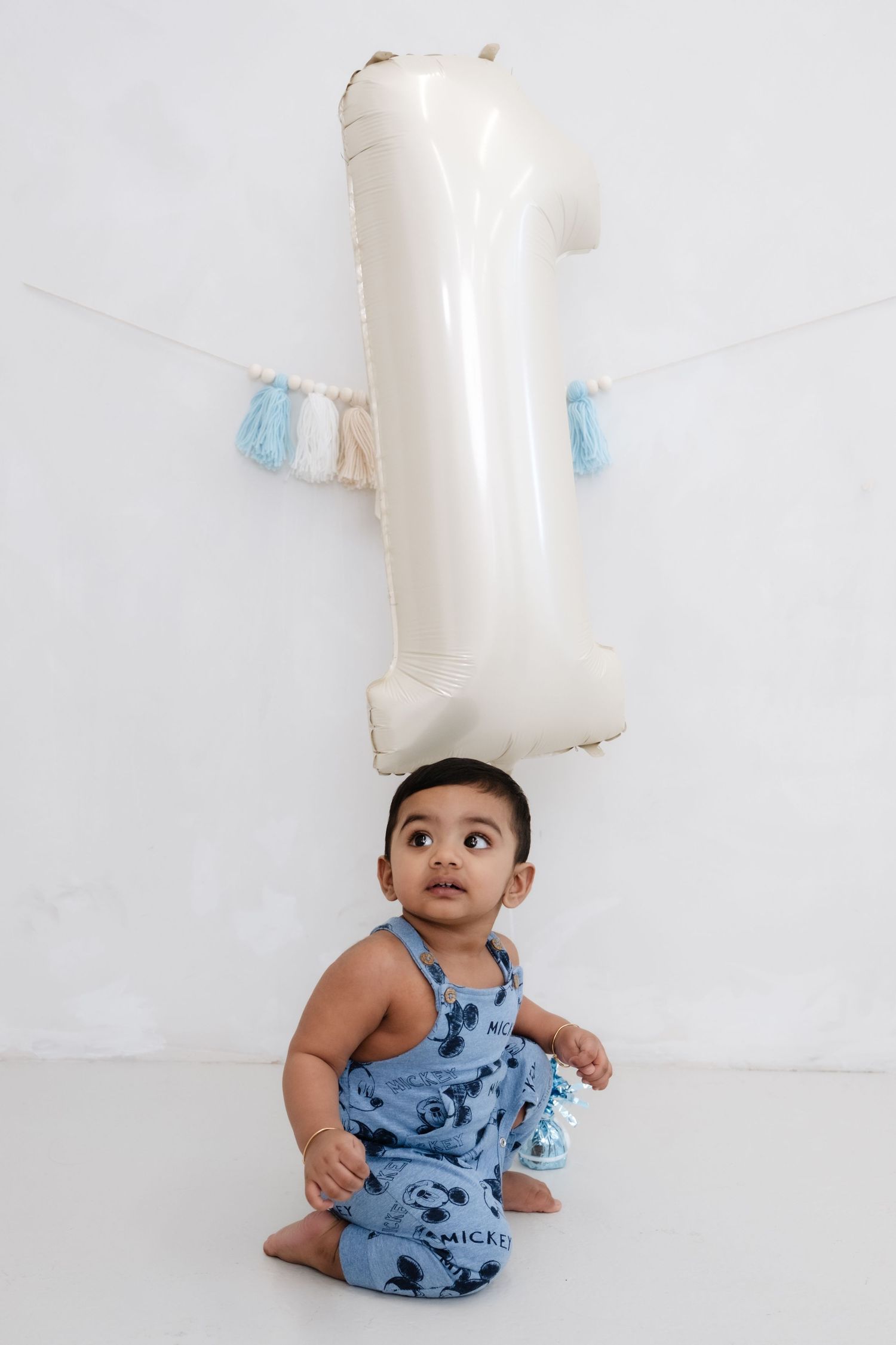 A baby in blue Mickey Mouse overalls sits on a white floor in front of a number 1 balloon for their first birthday.
