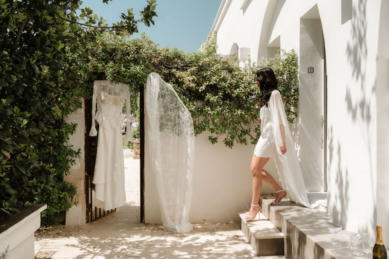 Woman in white dress walks up stone steps beside flowing white curtains and lush greenery at a sunlit Mediterranean villa.