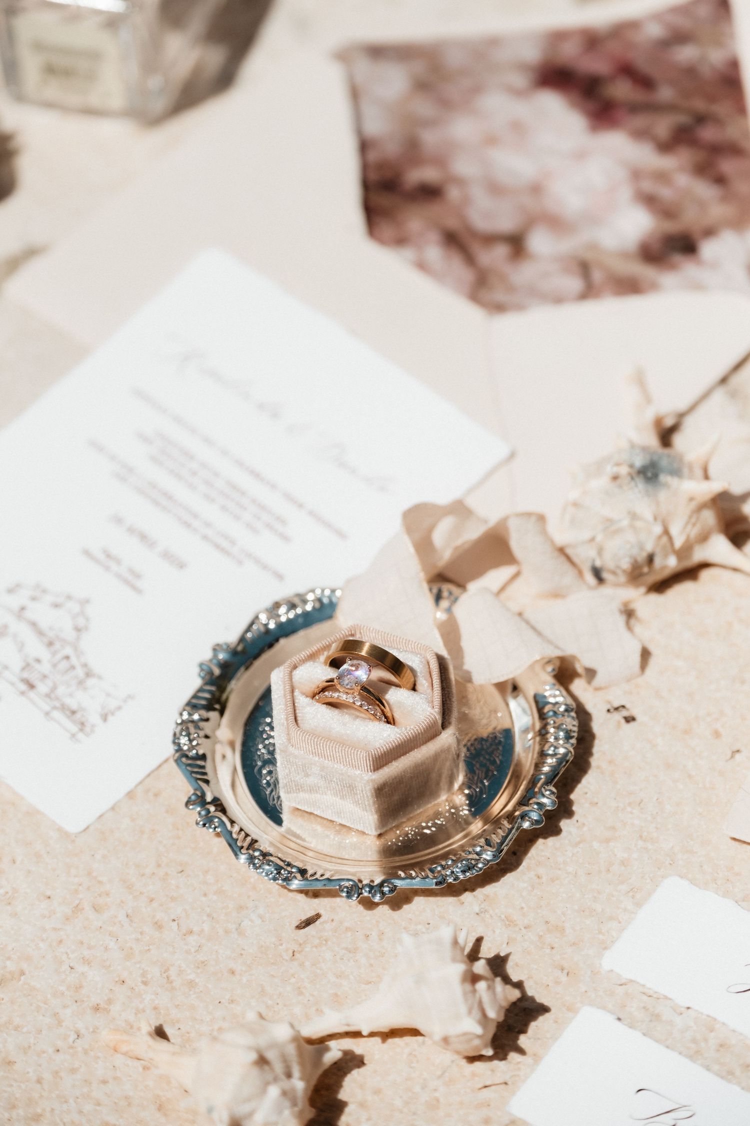 Gold engagement ring in a velvet hexagon box on a silver tray, surrounded by wedding stationery and dried florals.