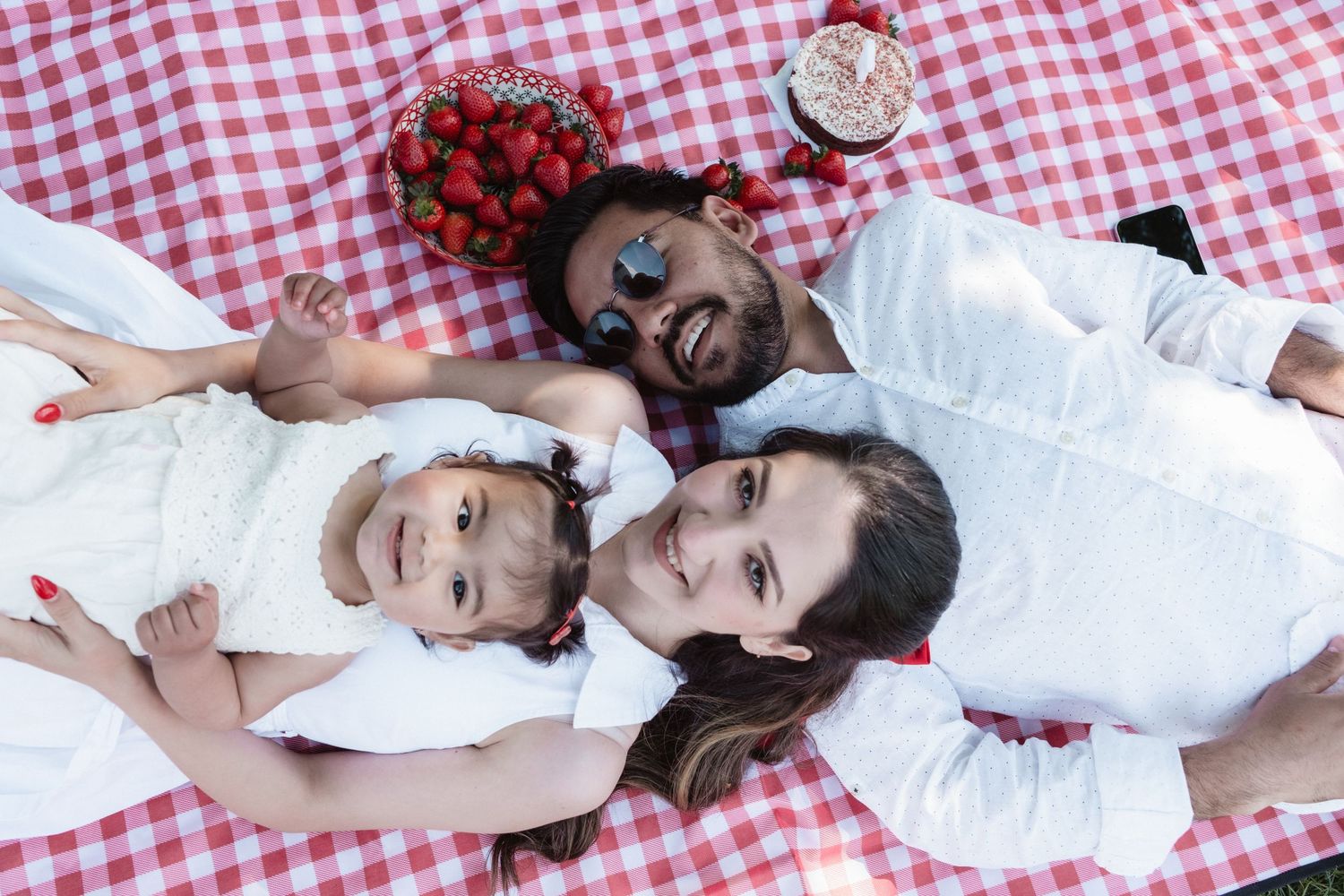 Family of three lying on red checkered picnic blanket with strawberries, smiling and enjoying a sunny outdoor picnic.