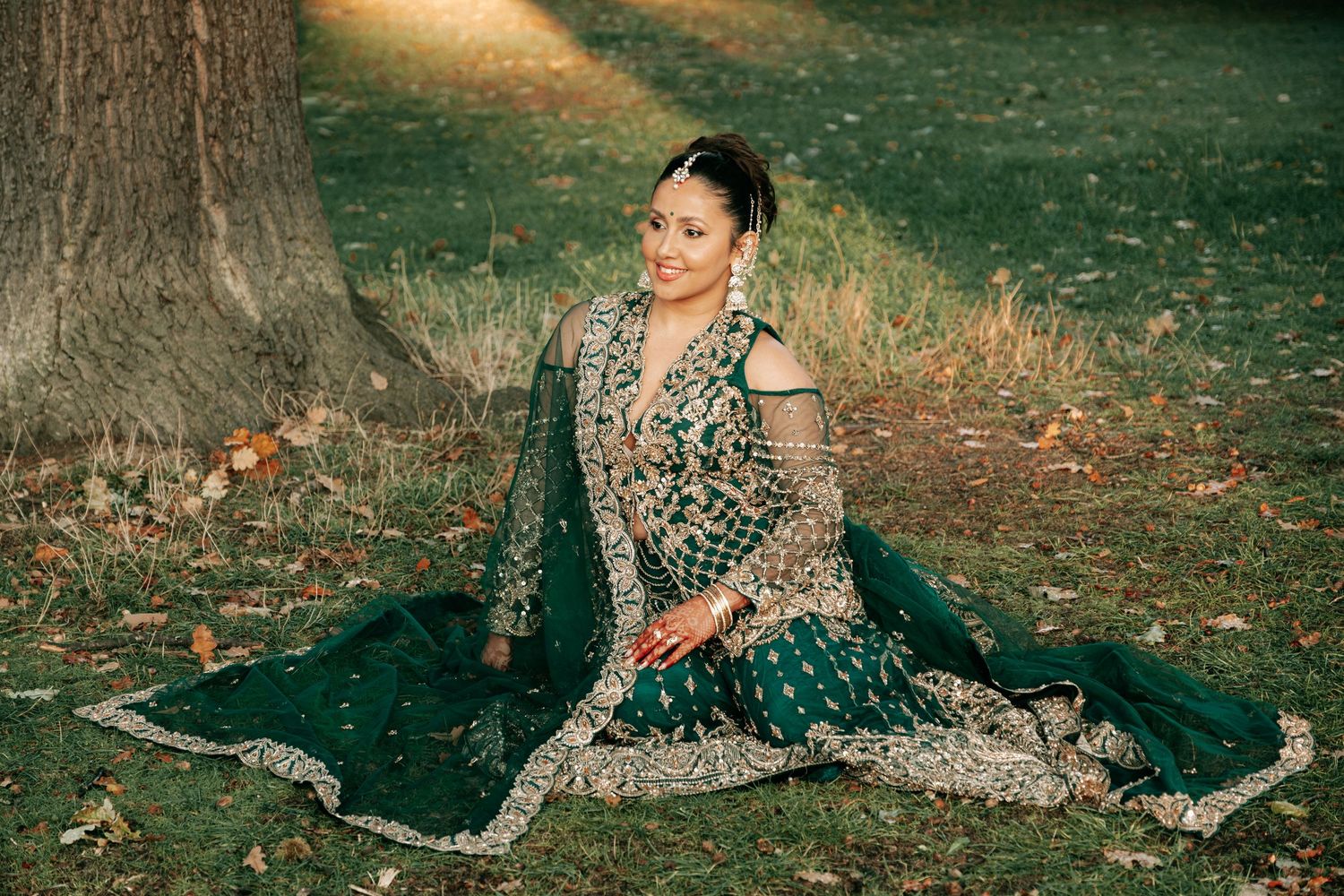 A bride in an ornate emerald green and gold lehenga poses gracefully on autumn leaves beneath a tree.
