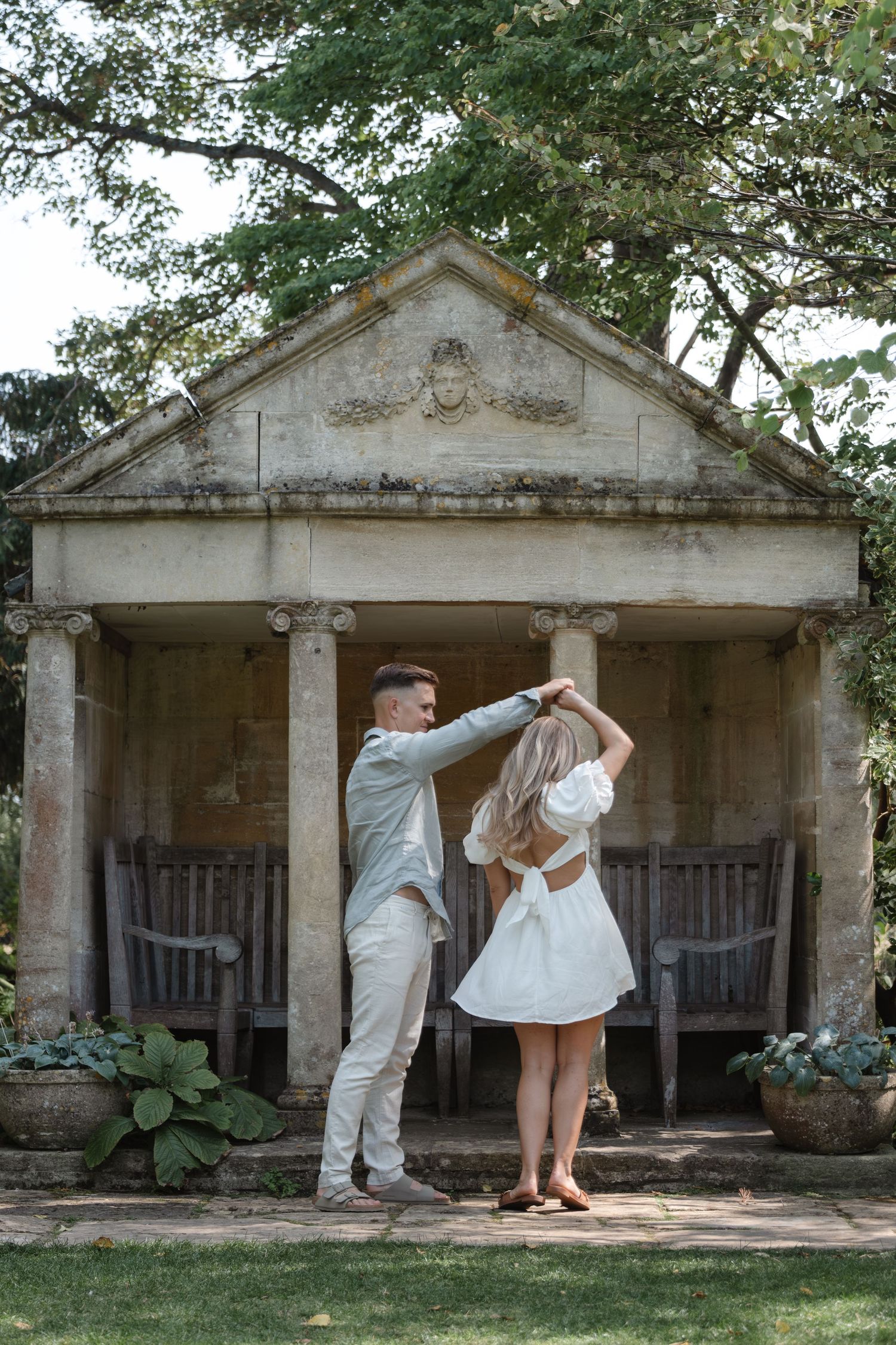 A couple poses romantically in front of a classical stone colonnade structure in a garden setting.