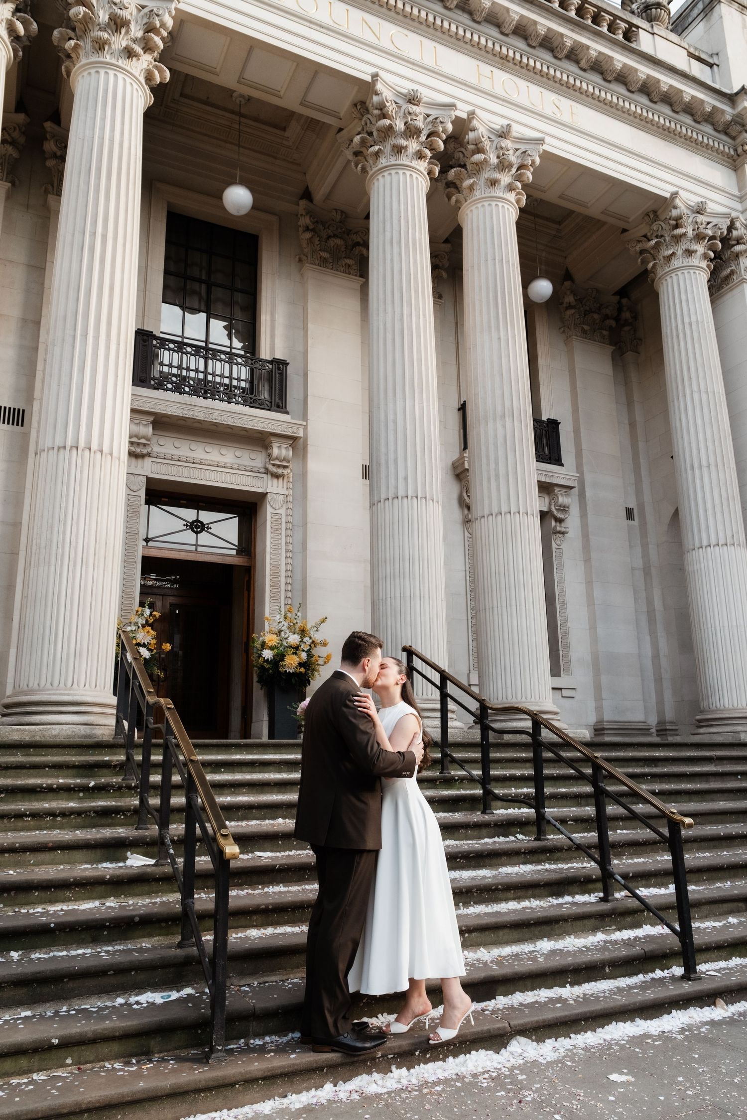 Newlyweds embrace on steps of grand neoclassical building with tall columns, bride in white dress, groom in brown suit.