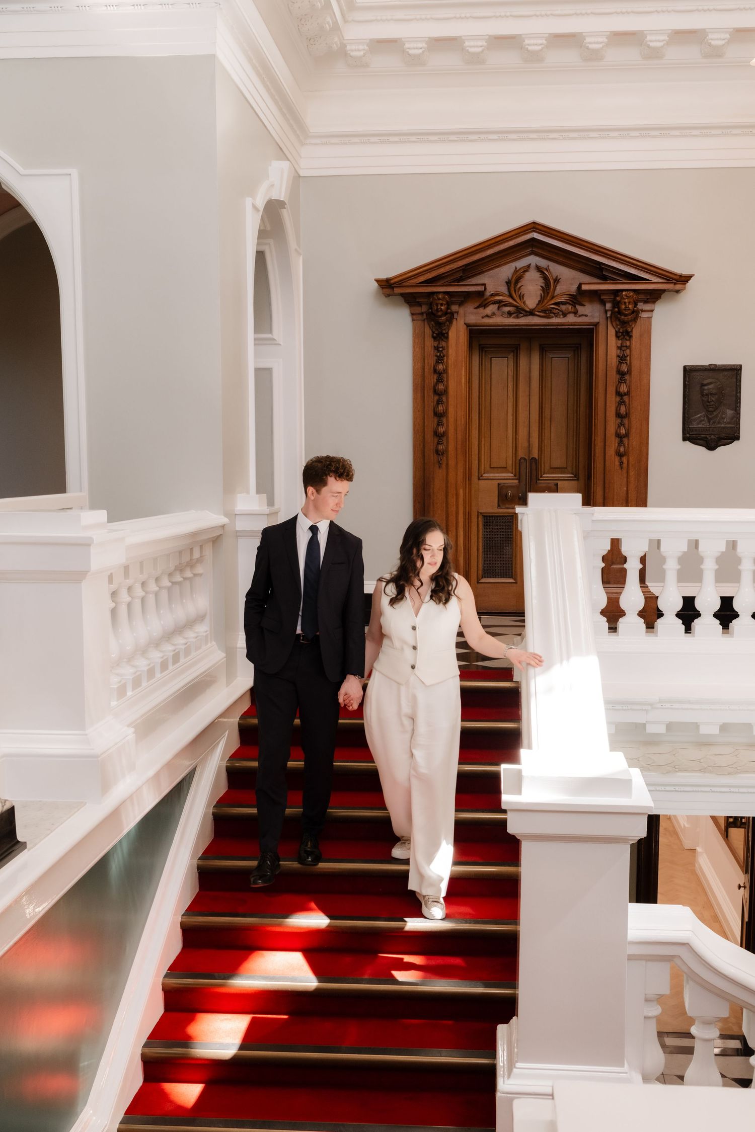 Elegant couple in formal attire walking down grand staircase with red carpet in a classic white interior.