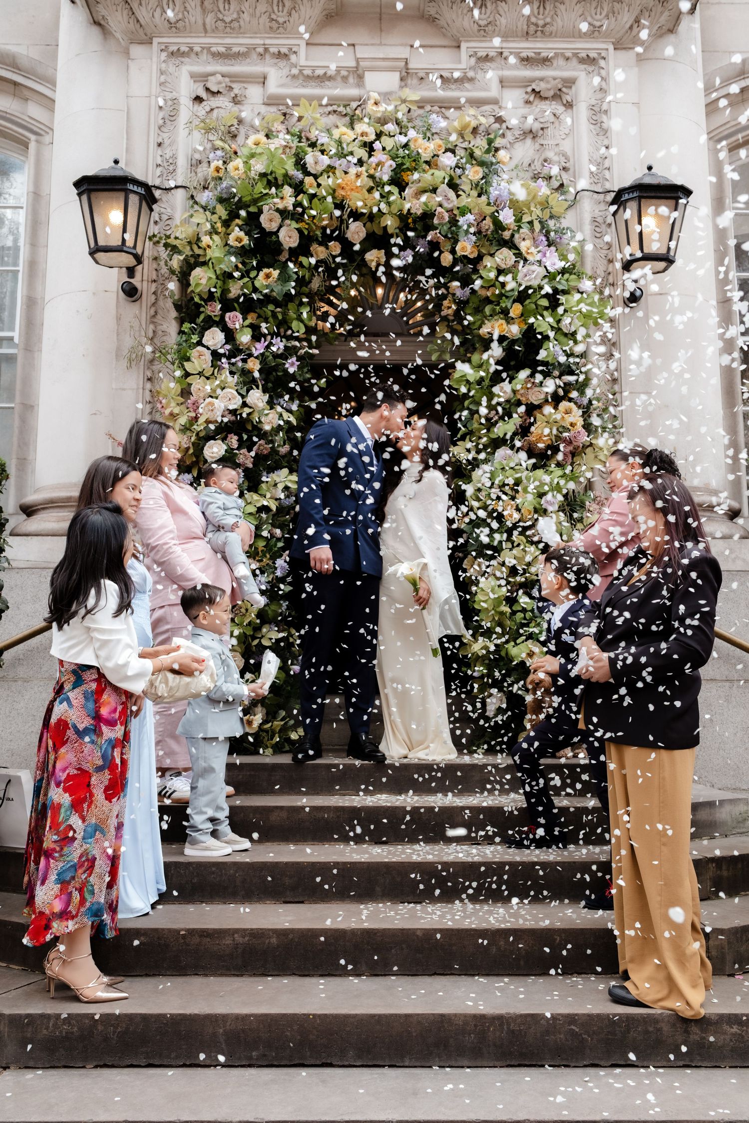 Couple kissing on steps surrounded by confetti and floral arch, with guests celebrating at an elegant wedding venue.