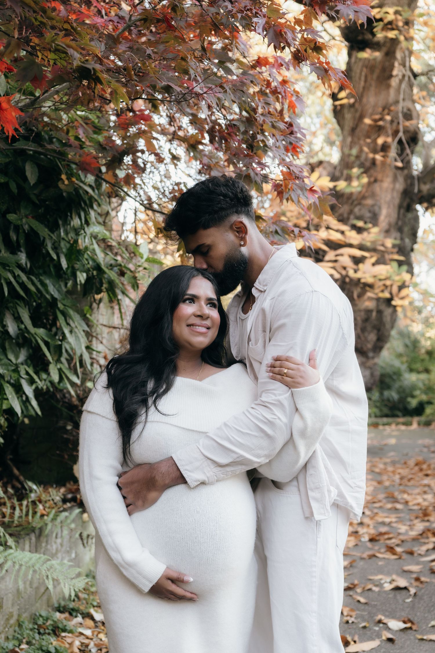 Maternity photo session in white outfits against autumn foliage backdrop.