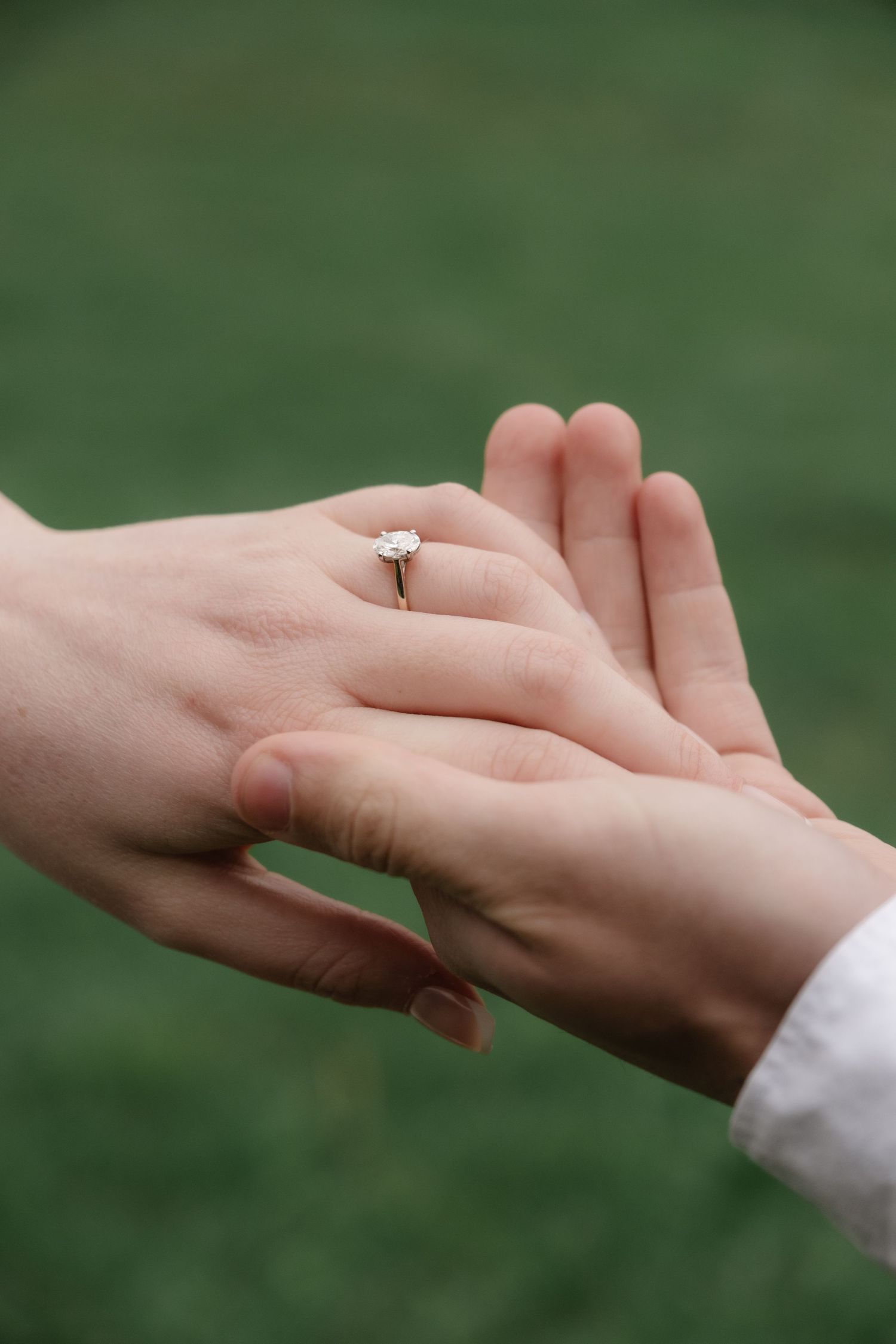 Close-up of two hands holding, one wearing a diamond solitaire engagement ring, with green grass background.