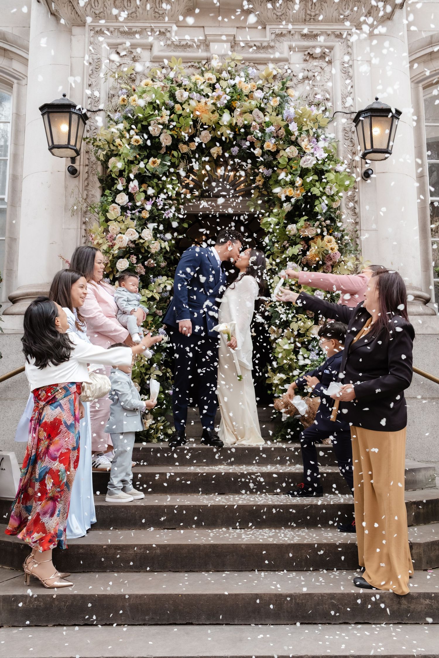 Bride and groom kiss on steps surrounded by confetti and floral arch, guests celebrating at elegant wedding venue.