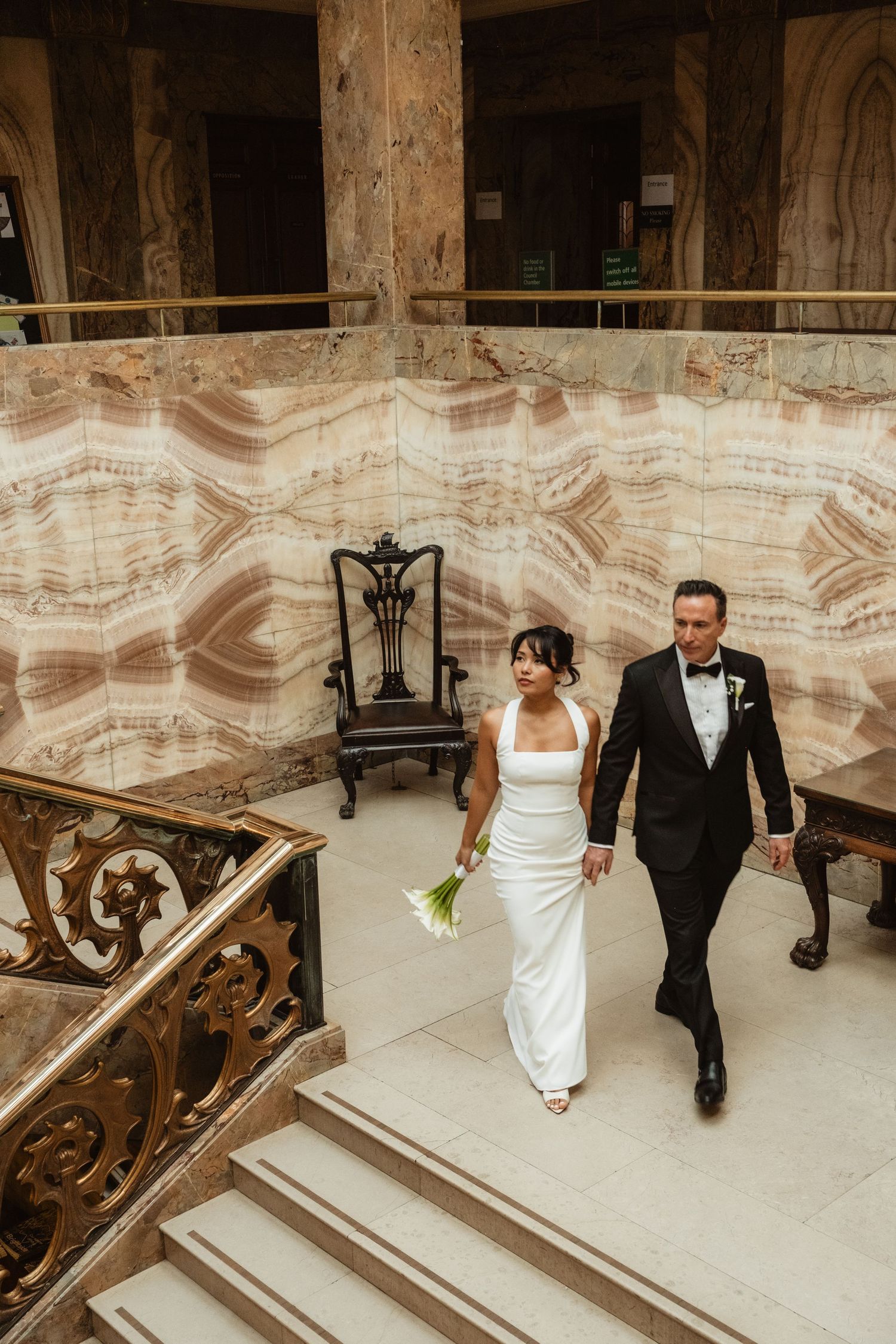 Couple walking down elegant marble staircase with ornate railings in formal wedding attire.