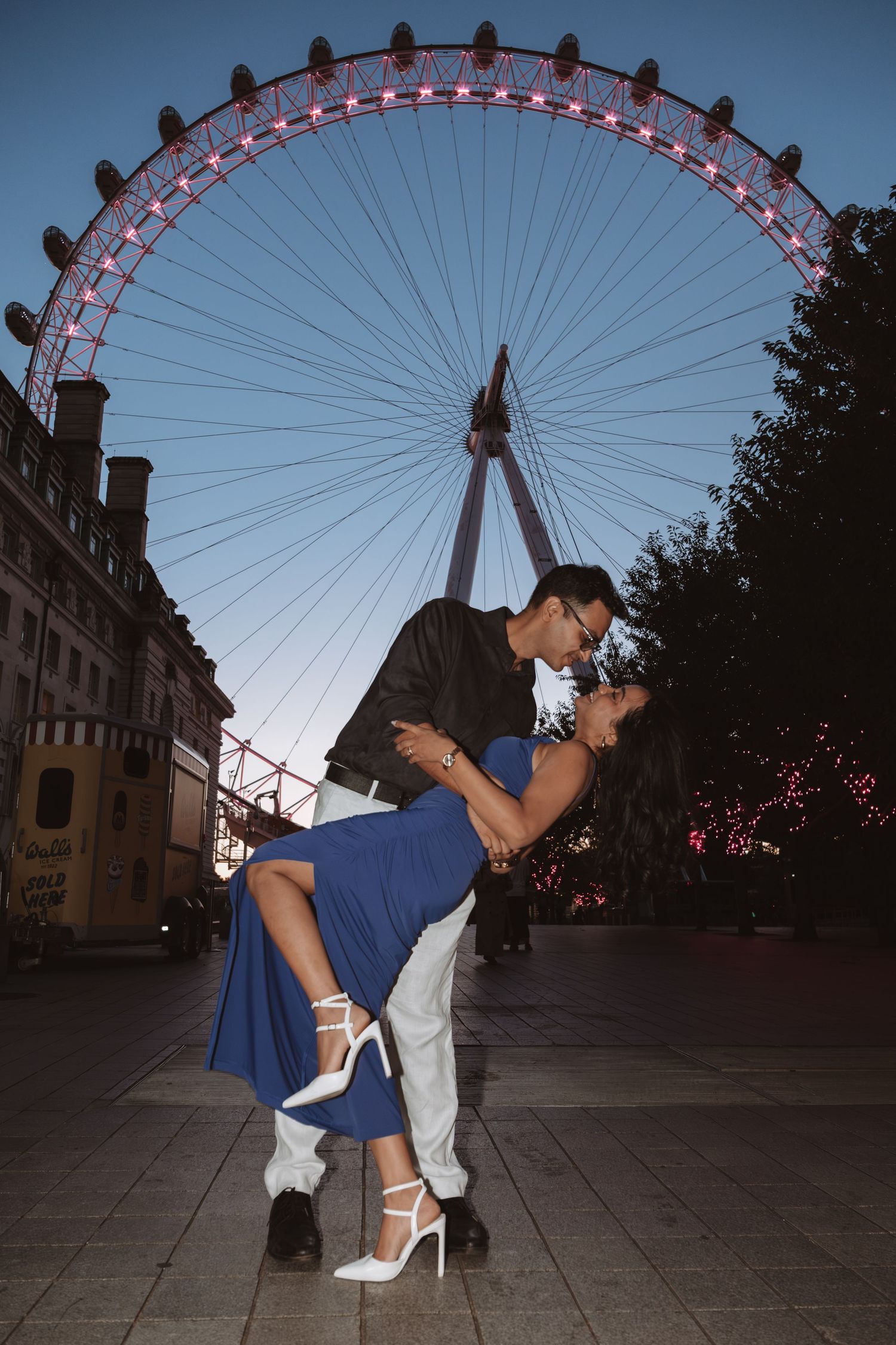A romantic dip pose with the illuminated London Eye ferris wheel creating a dramatic backdrop at dusk.