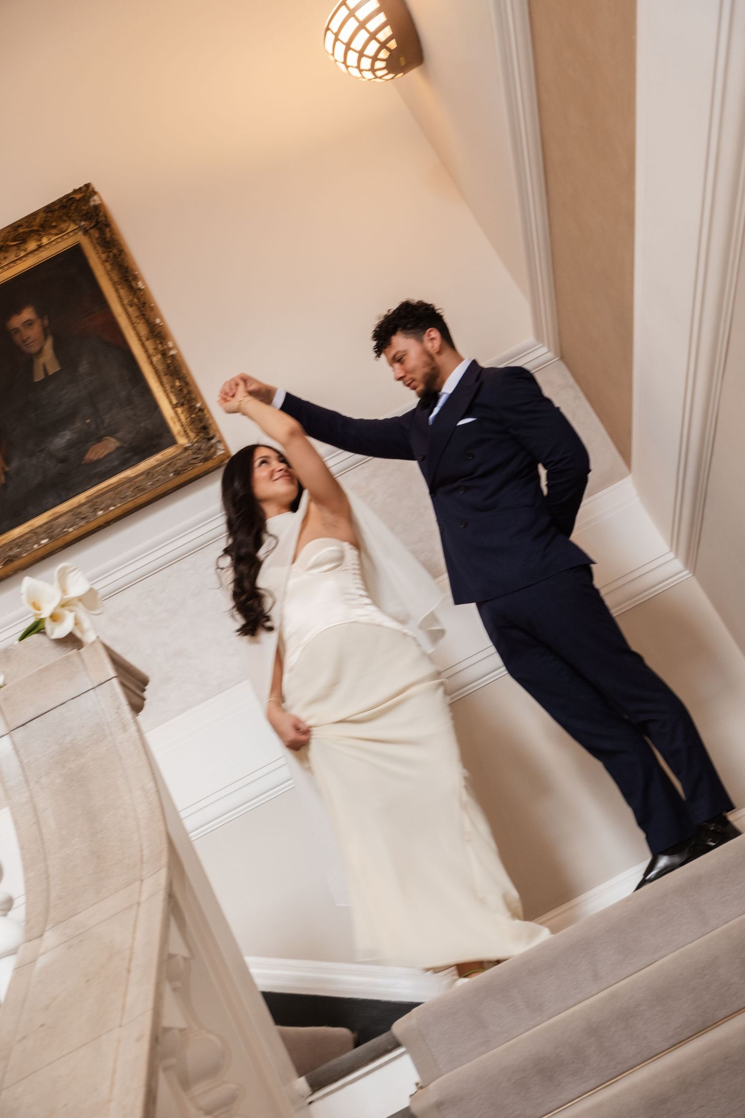 Bride and groom dancing elegantly on marble stairs, she in white gown, he in navy suit, inside a grand venue.