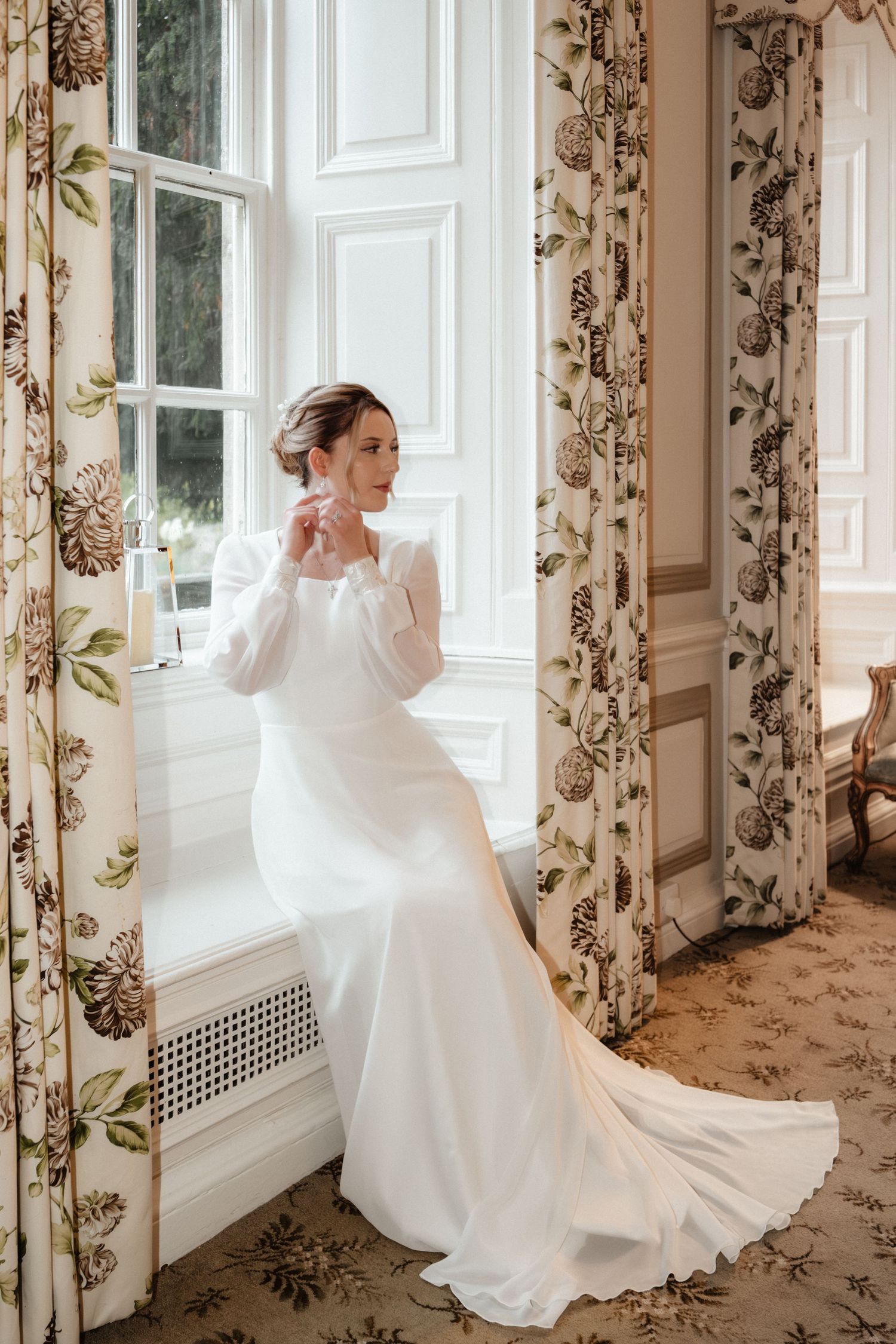 Elegant bride in flowing white gown poses by window with floral curtains.