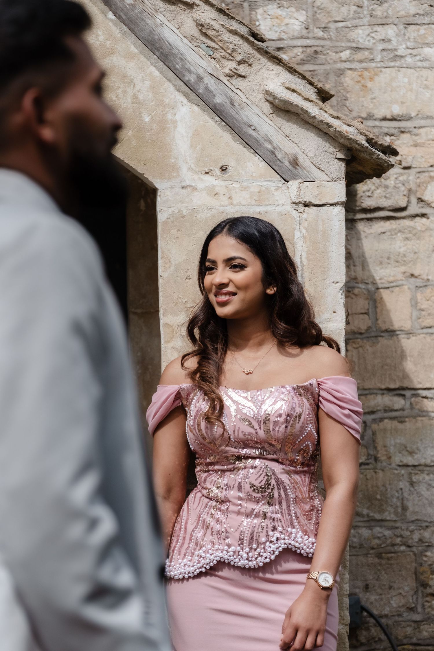 Woman in pink off-shoulder sequin gown smiling at man in grey suit near stone wall.