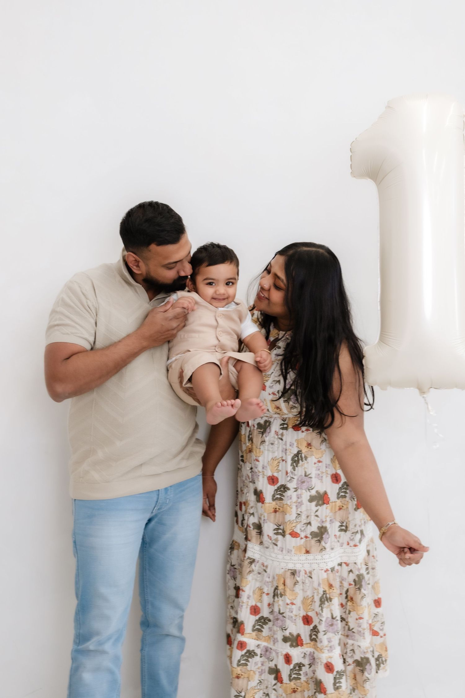 A family of three celebrates baby's first birthday, parents kissing their infant near a number one balloon.