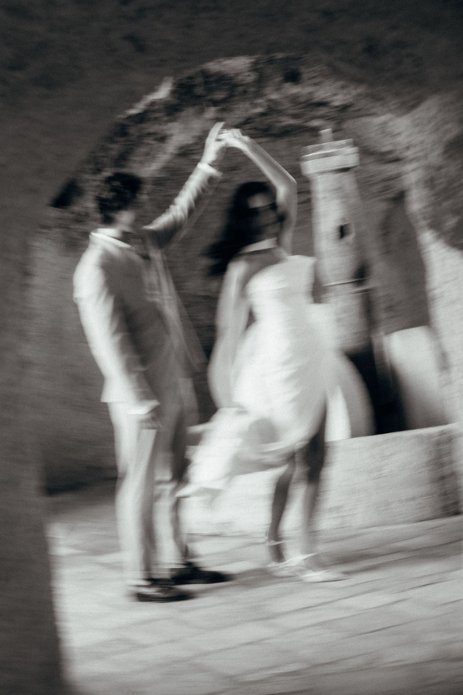 Blurred black and white photo of a bride and groom dancing near a stone fountain under an archway.