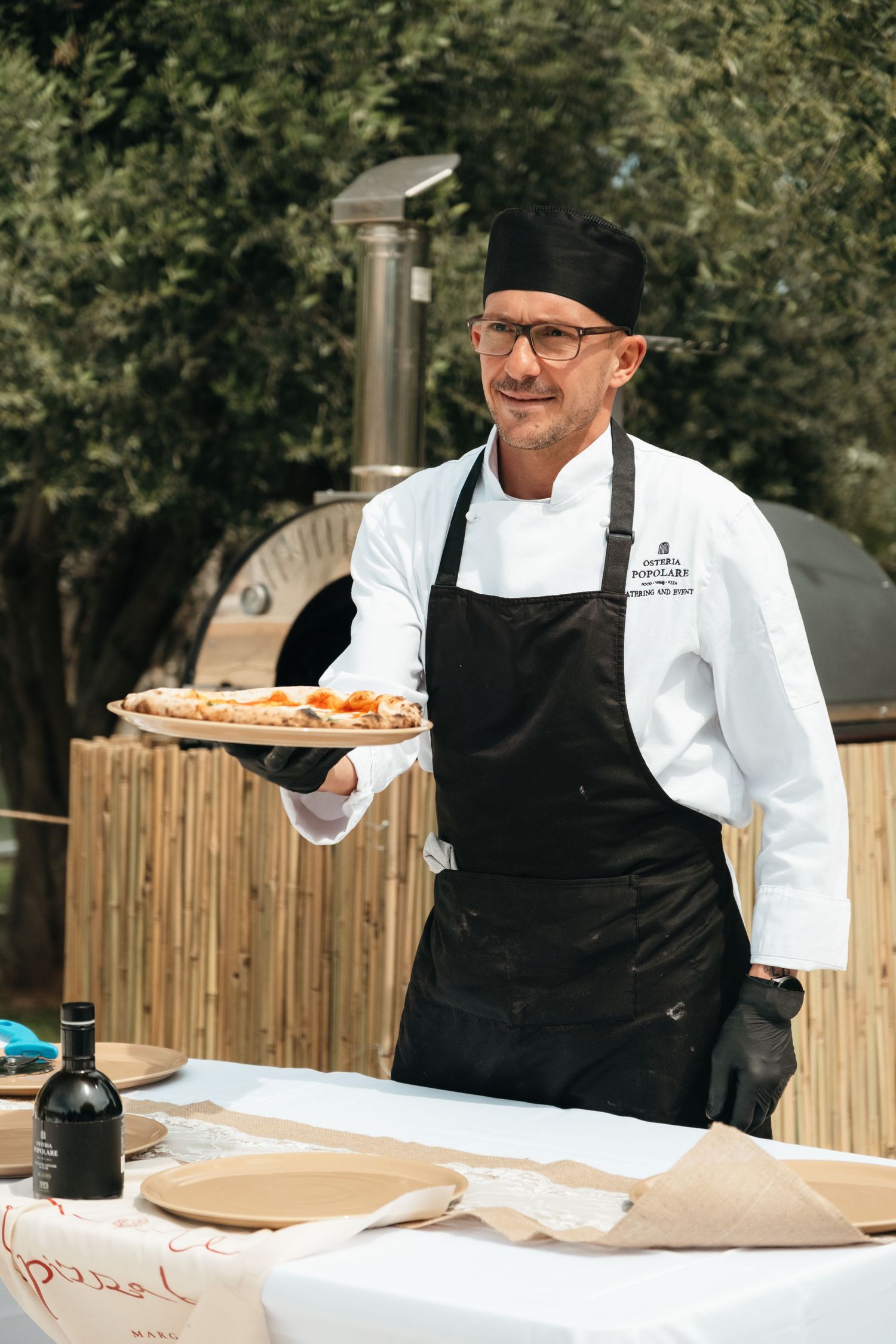 Chef in white coat and black apron proudly presents a freshly baked pizza at an outdoor wood-fired oven.