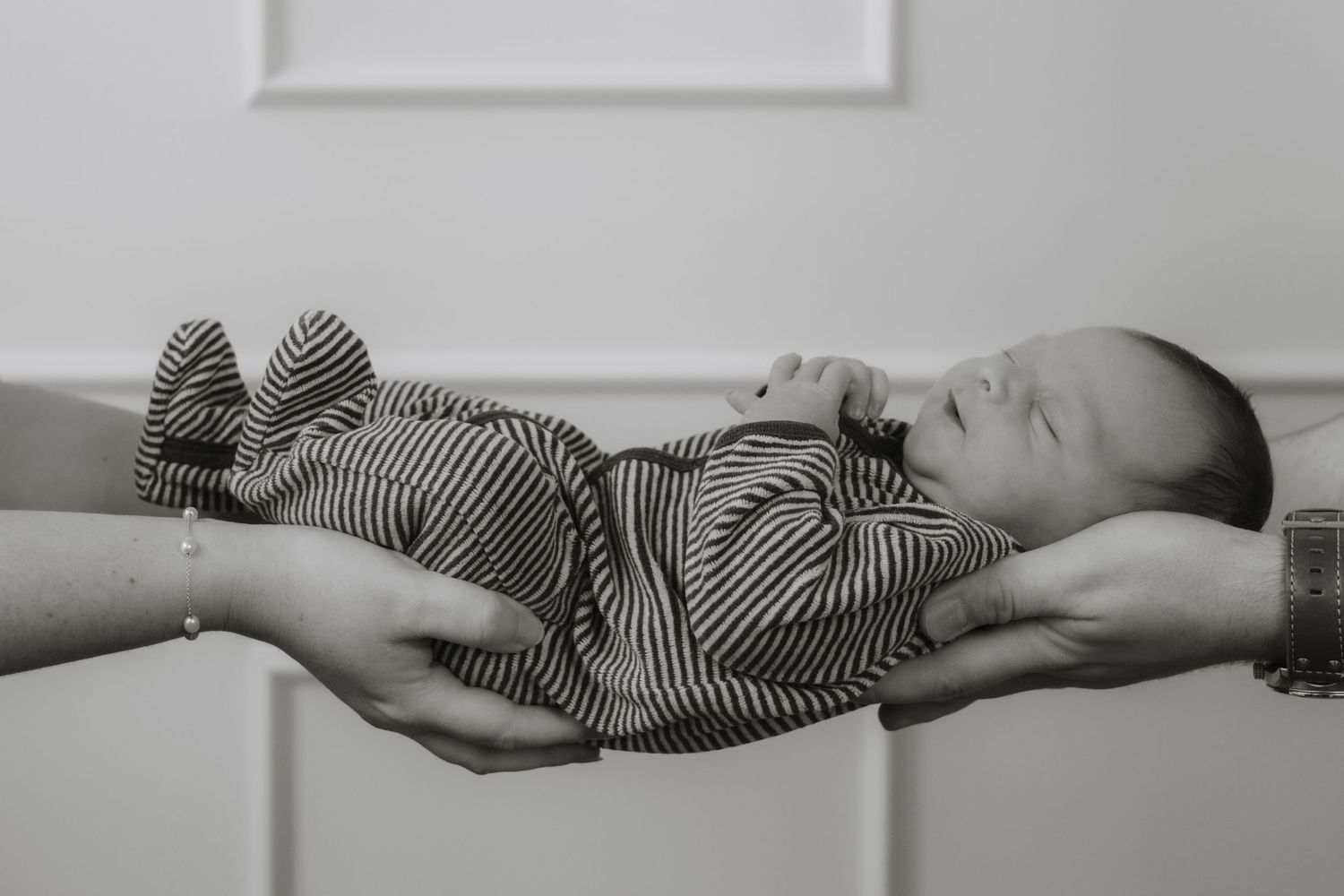 A sleeping newborn in striped outfit being gently cradled by adult hands.