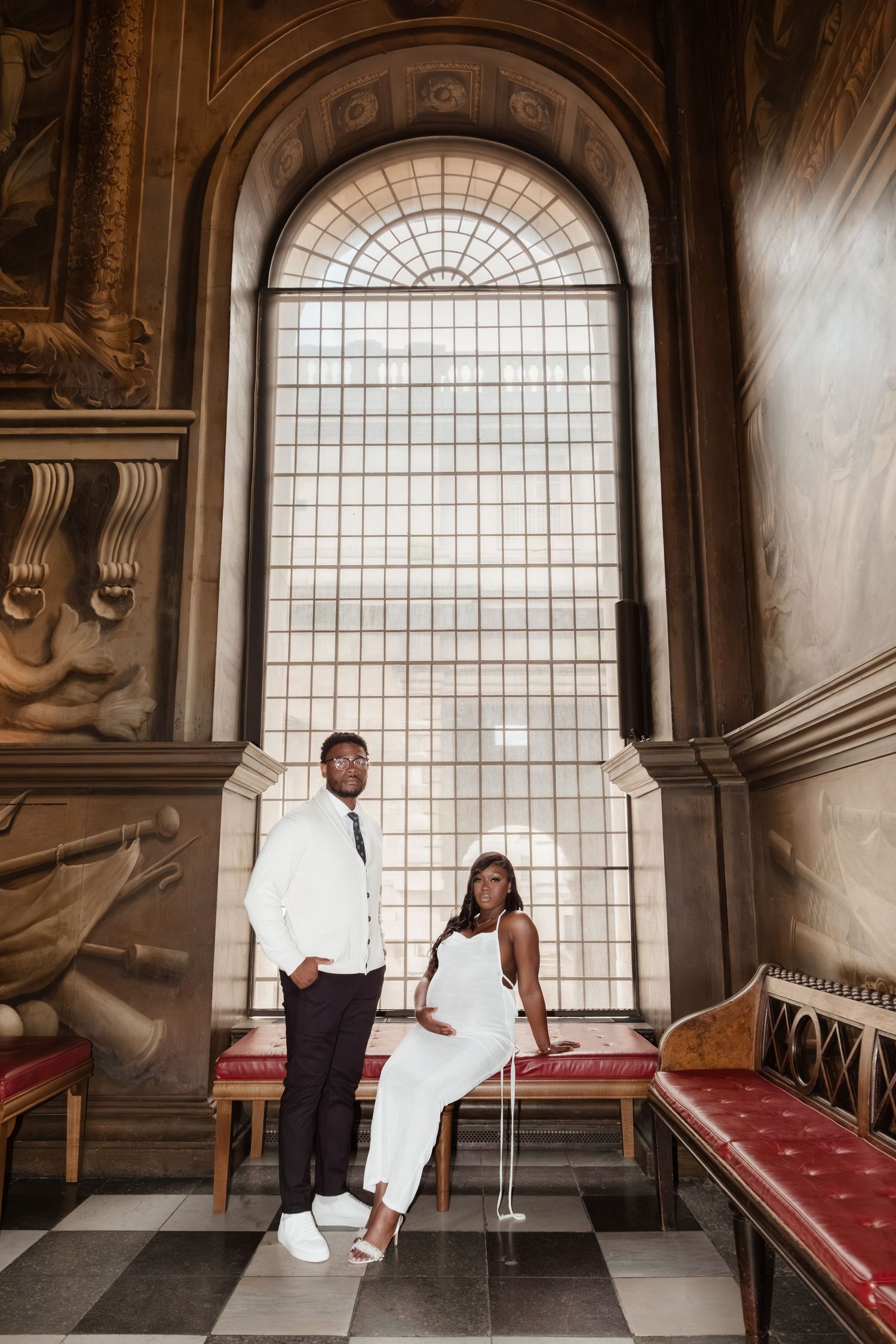 A couple poses in a grand historic hall with classical columns and large arched windows.