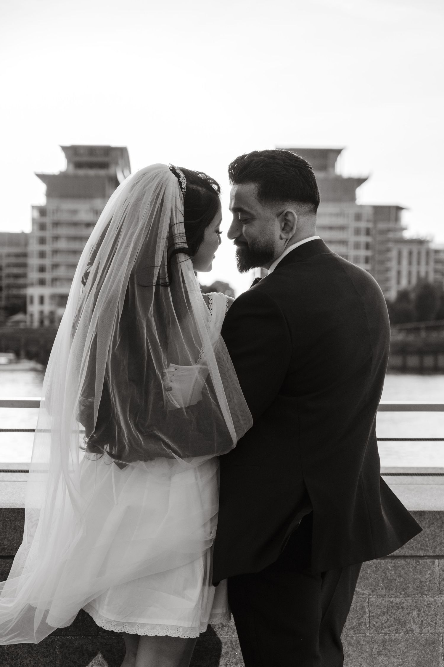 A romantic black and white wedding portrait of a couple by waterfront with city buildings in background.