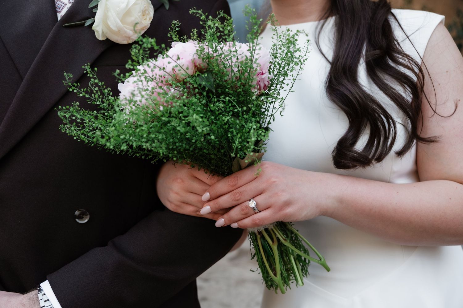 Bride and groom holding a wedding bouquet of pink and white flowers with greenery, bride wearing diamond engagement ring.