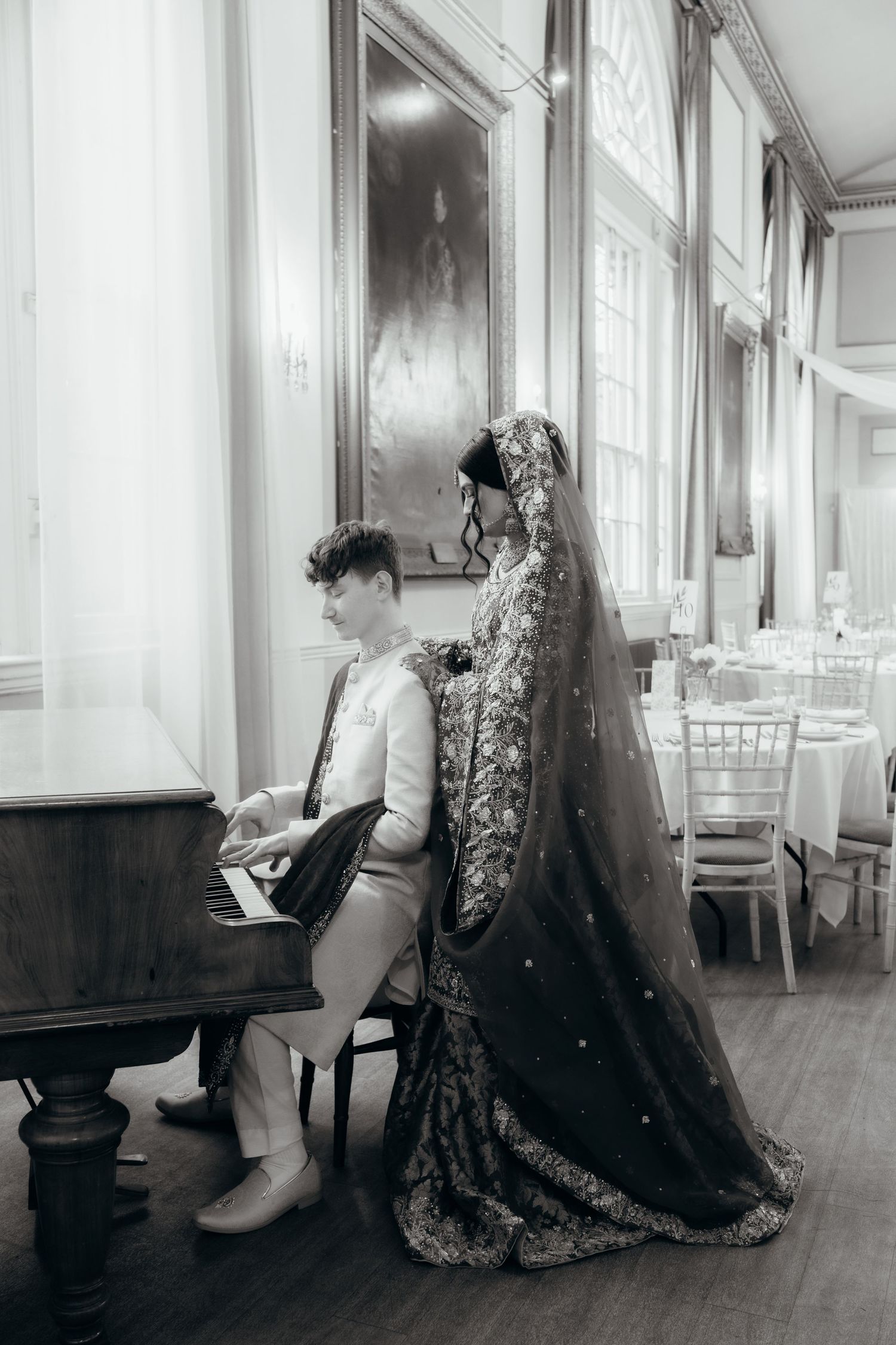 Black and white photo of a groom playing piano while bride in ornate gown and veil stands beside him in elegant venue.