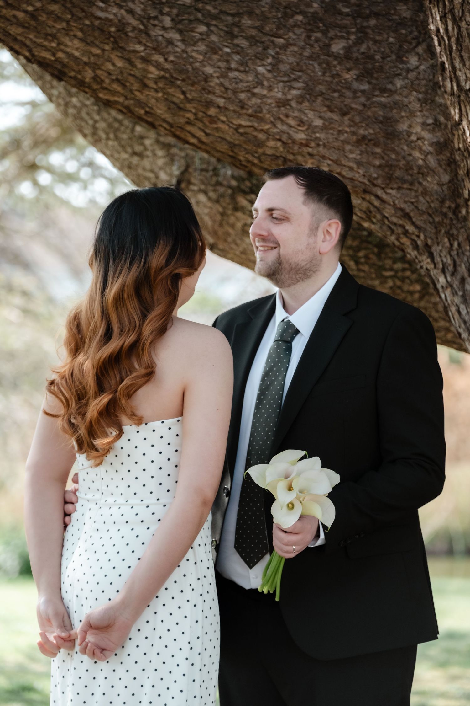 Smiling groom in black suit holds white calla lily bouquet beside bride in polka dot dress under a tree.