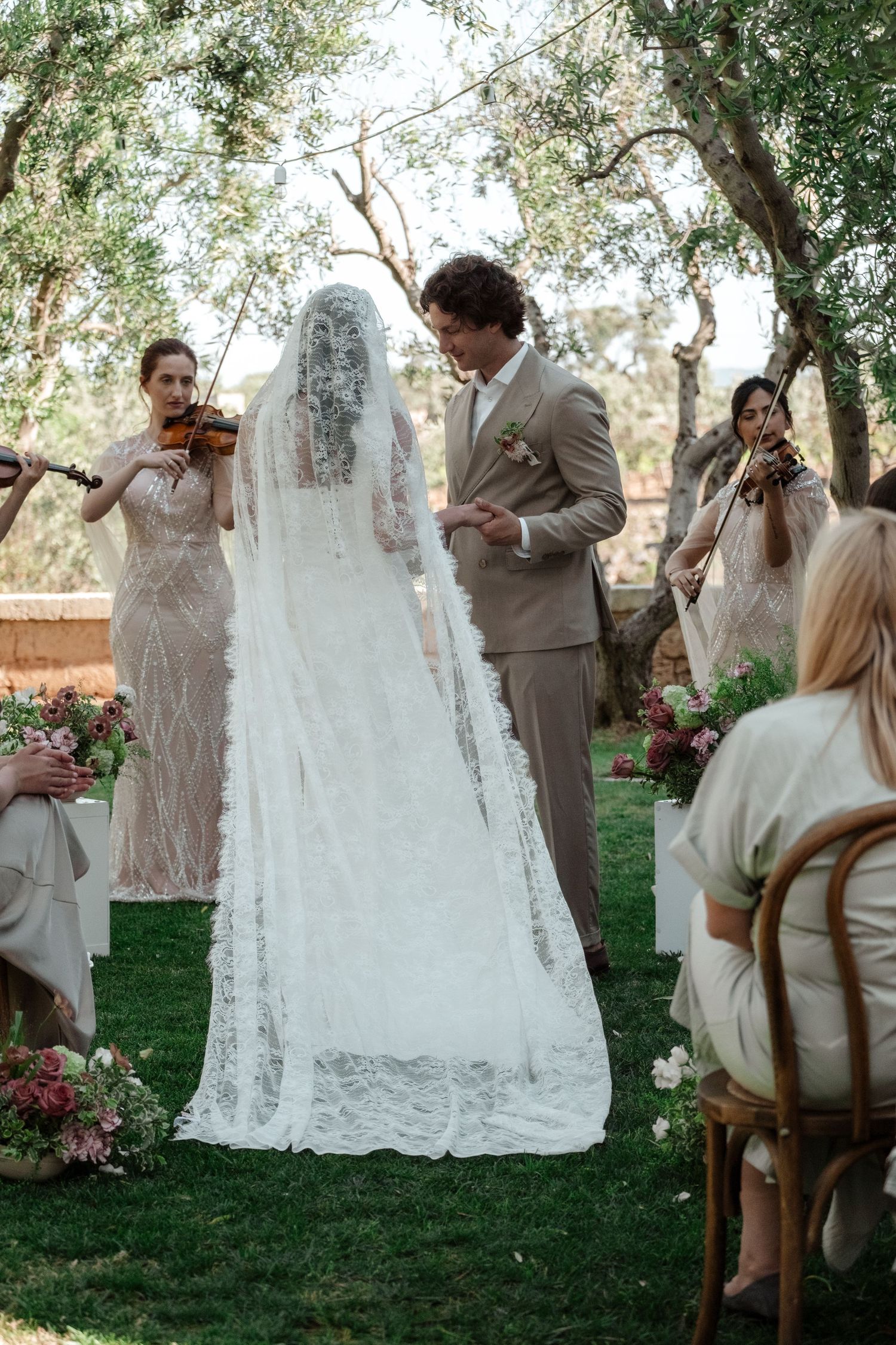 Bride and groom exchange vows outdoors under olive trees with violinists playing during an elegant garden wedding ceremony.