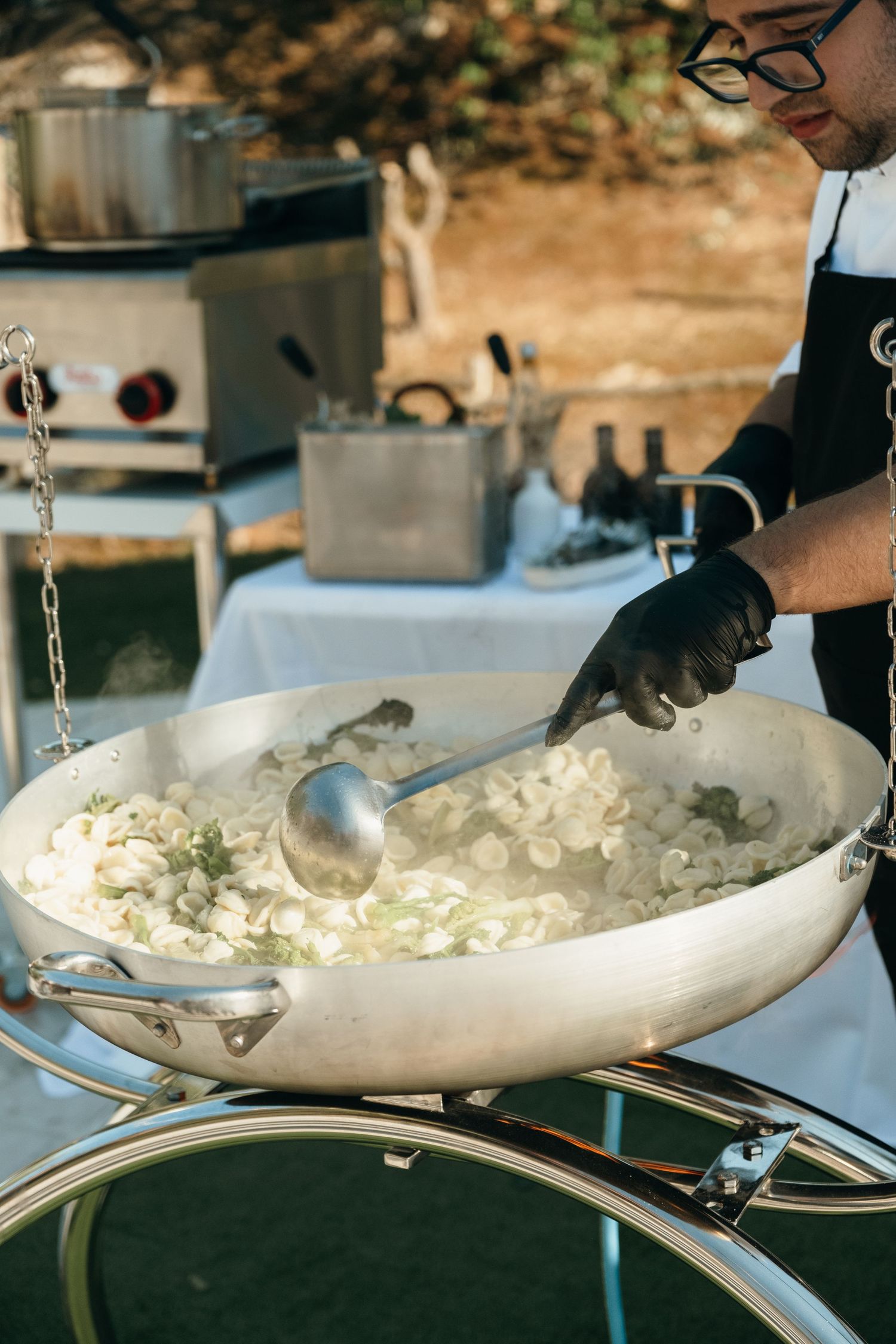 Chef in black gloves stirring vegetables in a large catering pan at an outdoor cooking event.